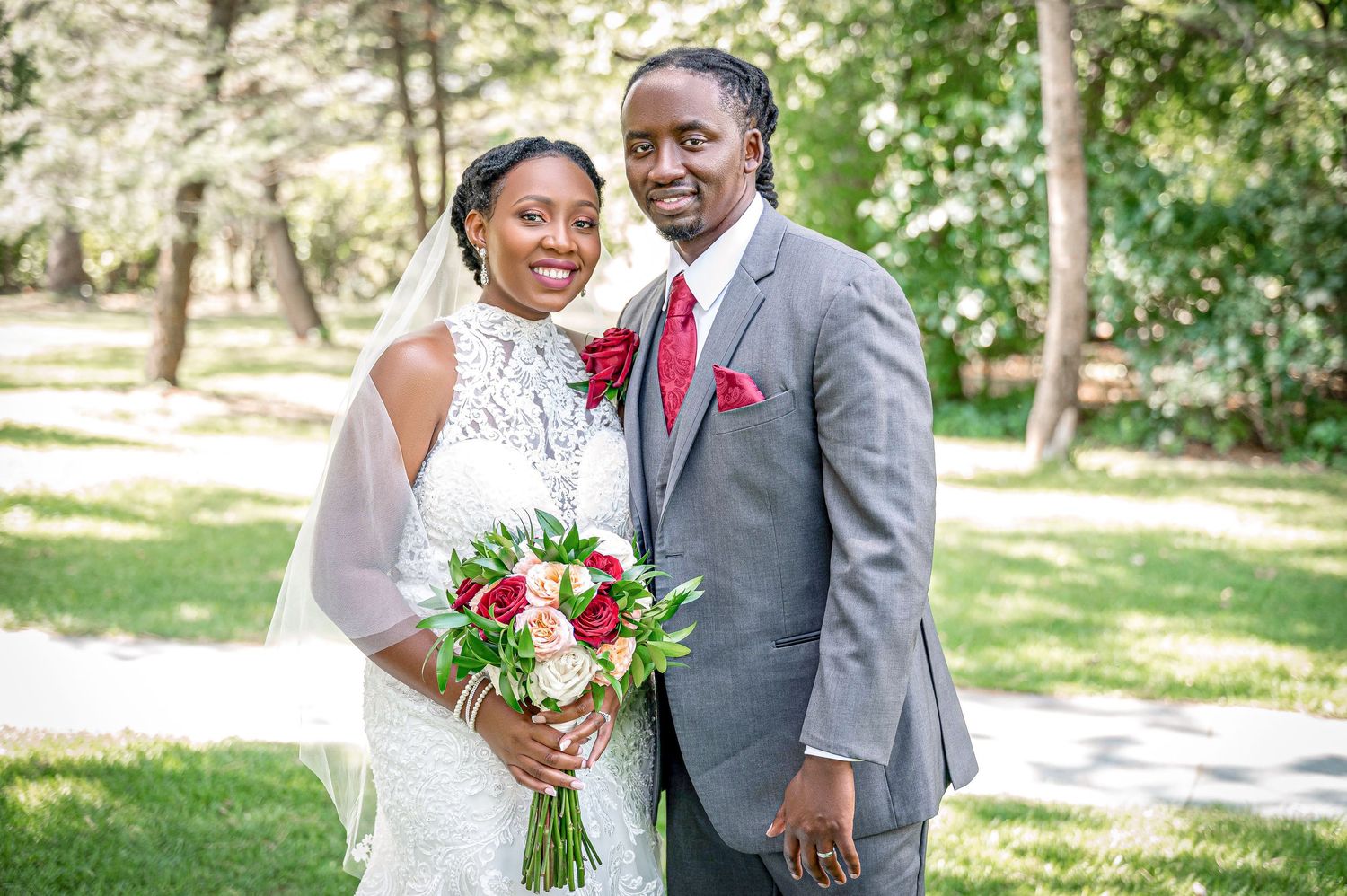 Newlyweds pose together outdoors, with bride holding red and white bouquet against natural backdrop.