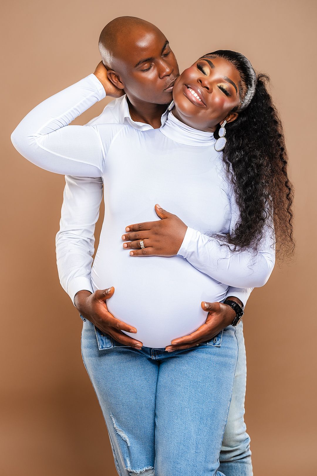 Intimate maternity photoshoot featuring couple in white top and jeans against beige background.