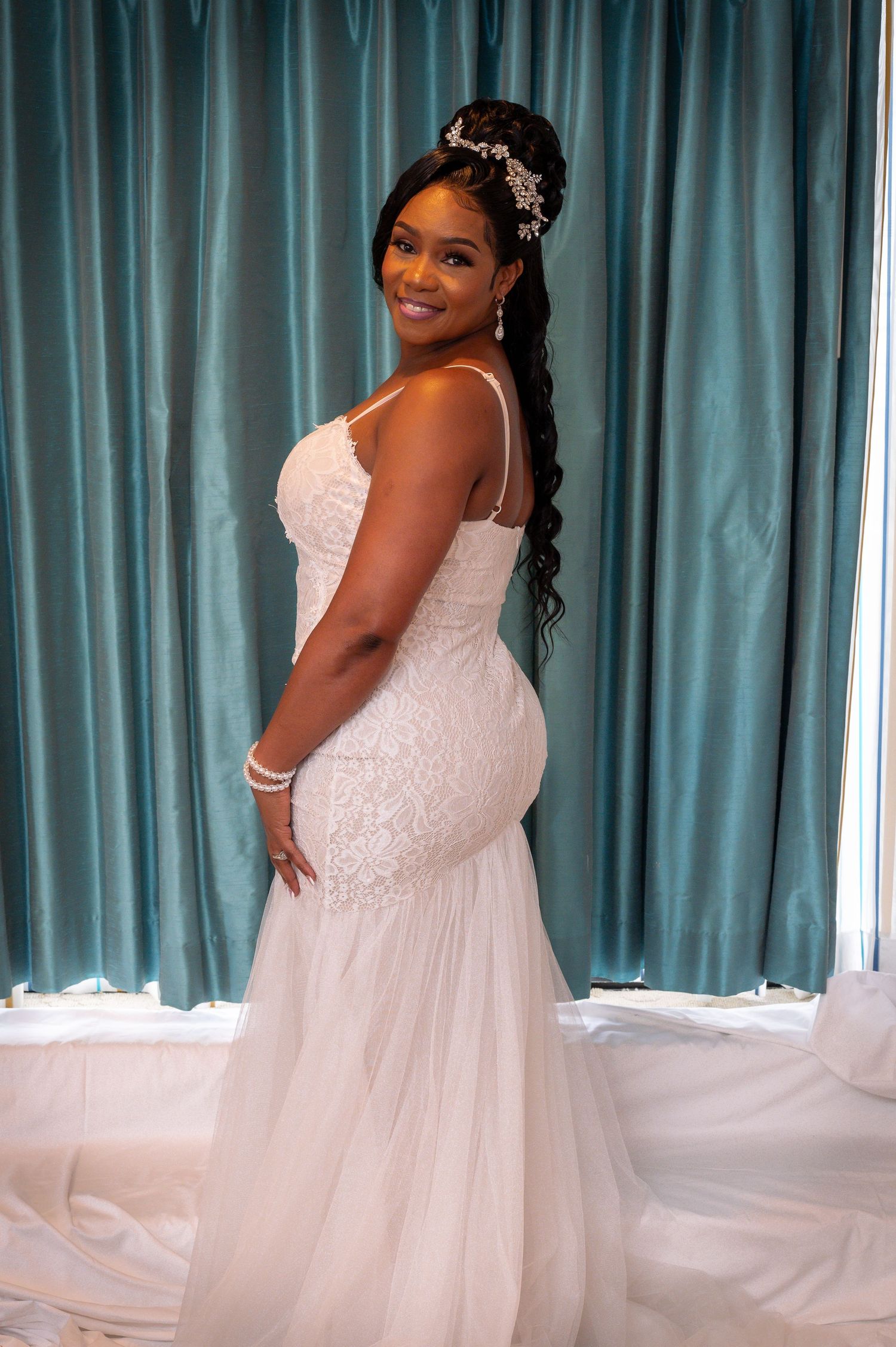 A person in a fitted lace wedding gown poses against a turquoise curtain backdrop.