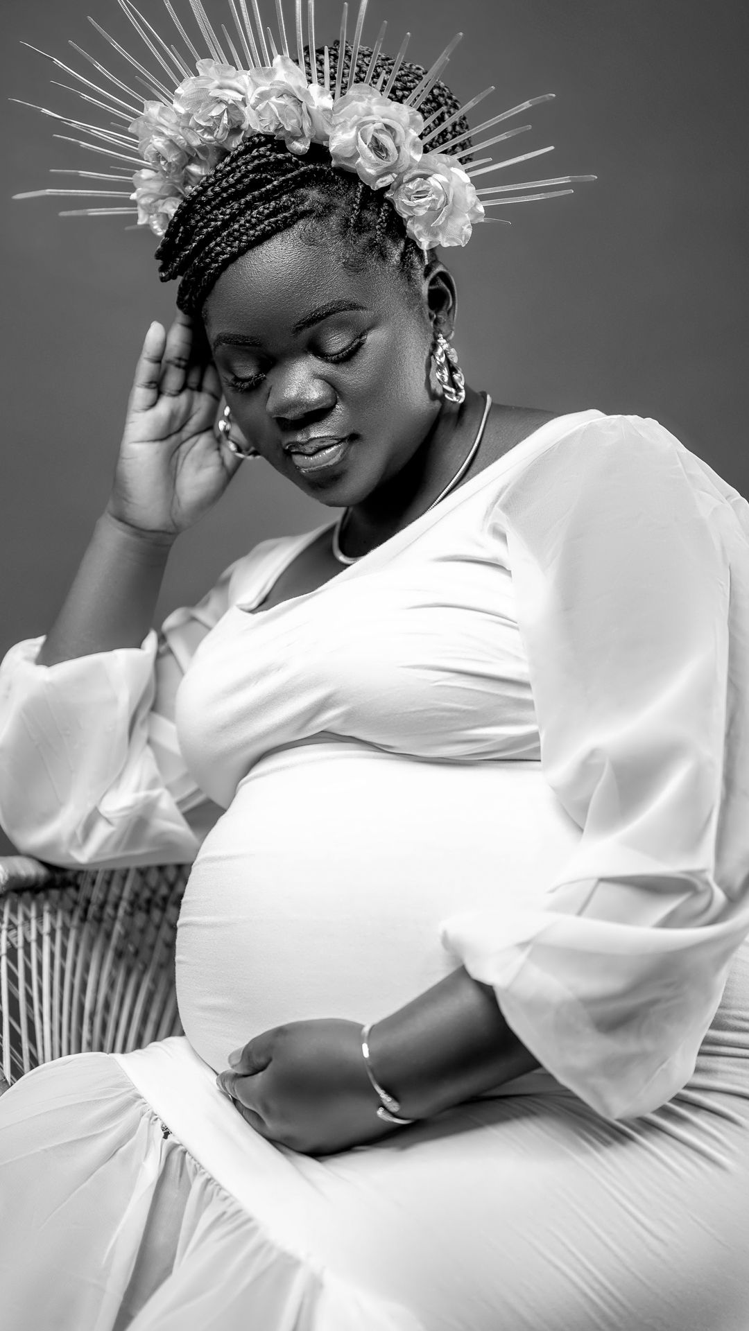 Black and white maternity photo featuring woman in white dress with floral crown seated elegantly.