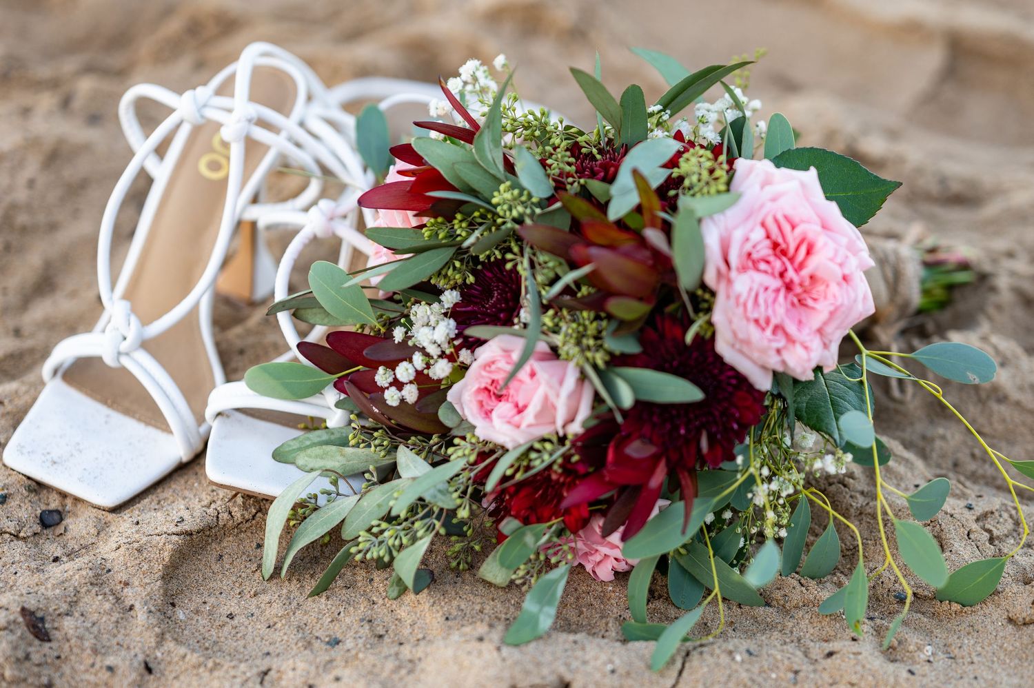 Wedding bouquet with pink garden roses and burgundy flowers laying beside white bridal shoes.