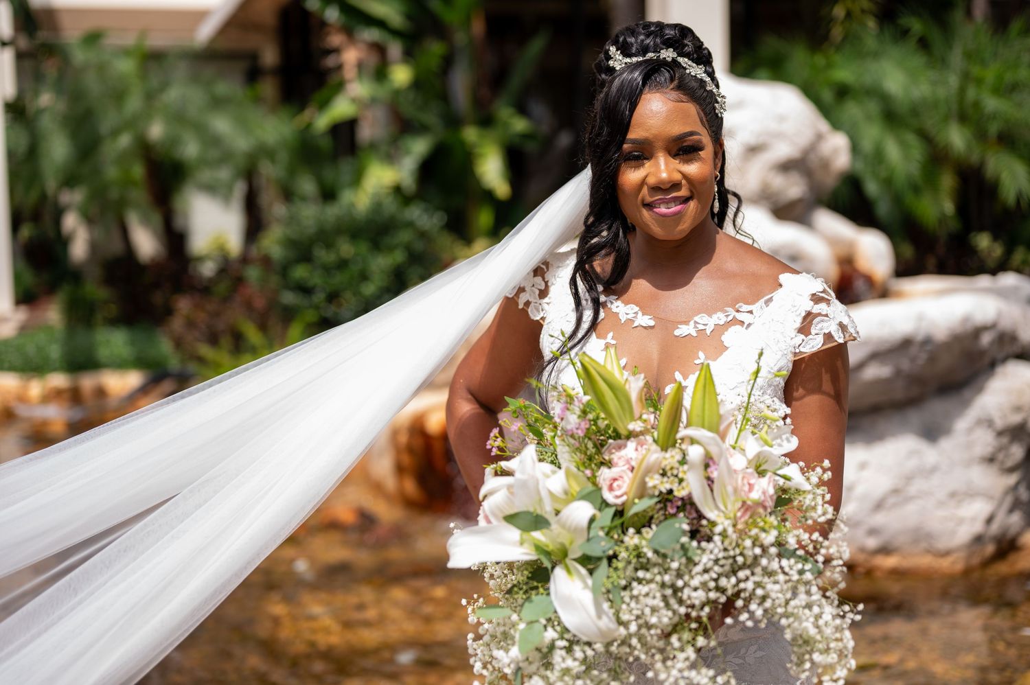 Bride in lace gown holds white floral bouquet against tropical garden backdrop.