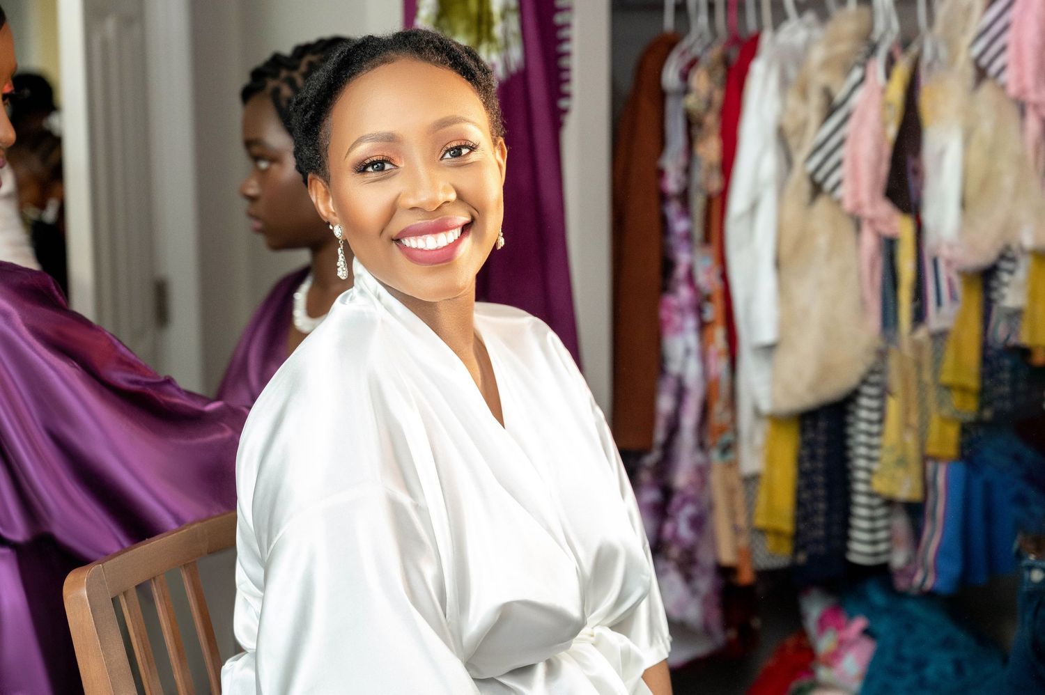 Woman in white robe smiles brightly in dressing room with clothing racks visible behind her.