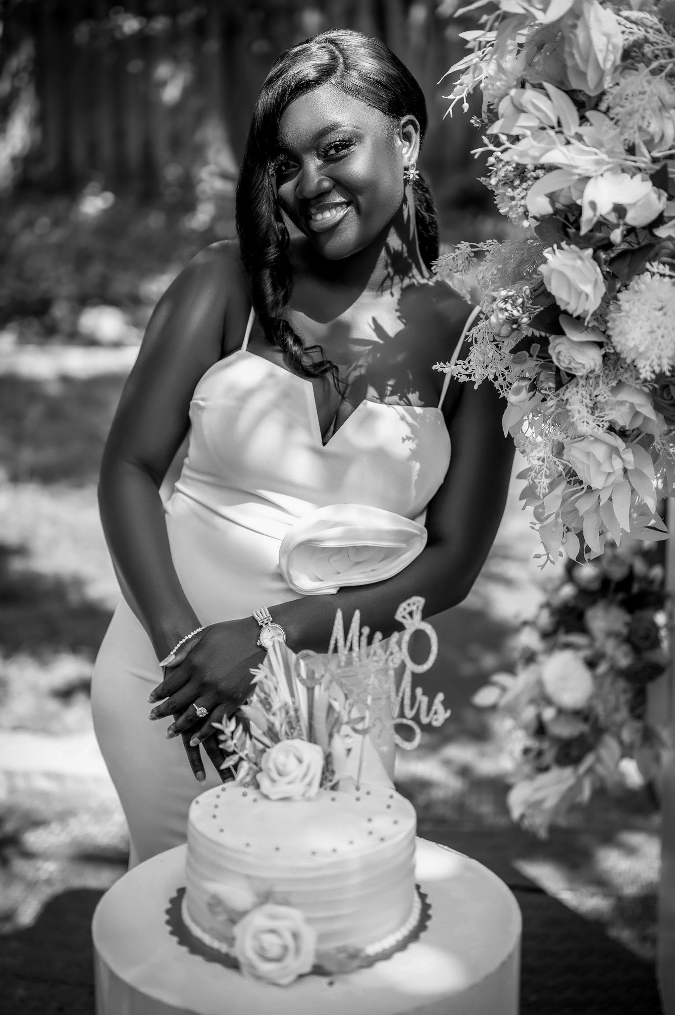 Black and white photo of bride in white dress smiling next to decorated wedding cake.
