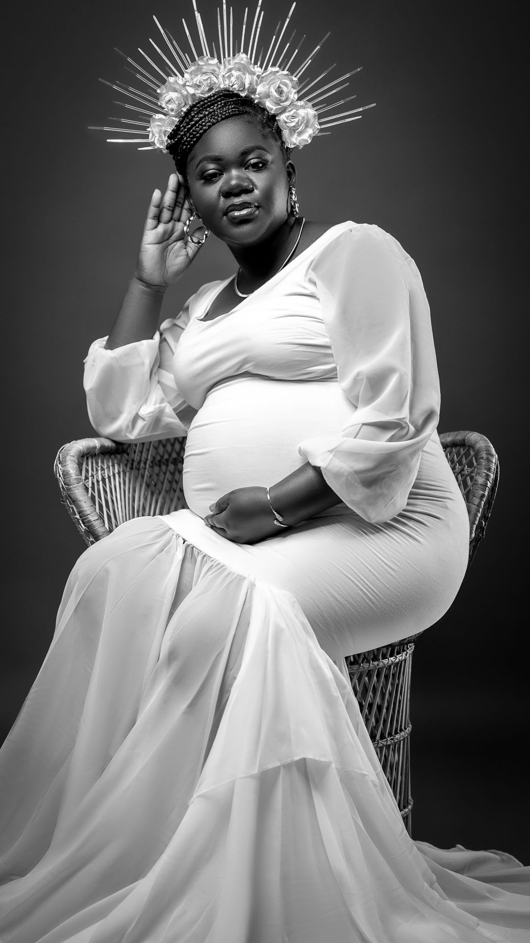 Maternity portrait in white dress with crown headpiece sitting in wicker chair against dark background.