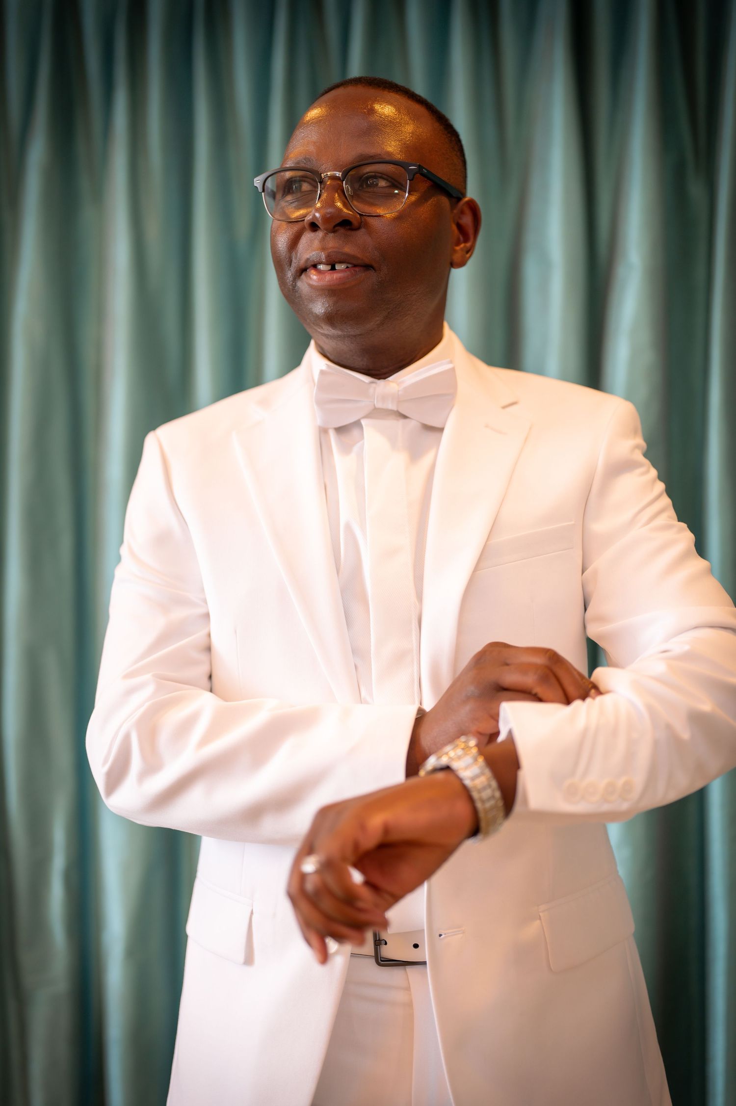 A person in a crisp white tuxedo and bowtie poses against a teal curtain backdrop.