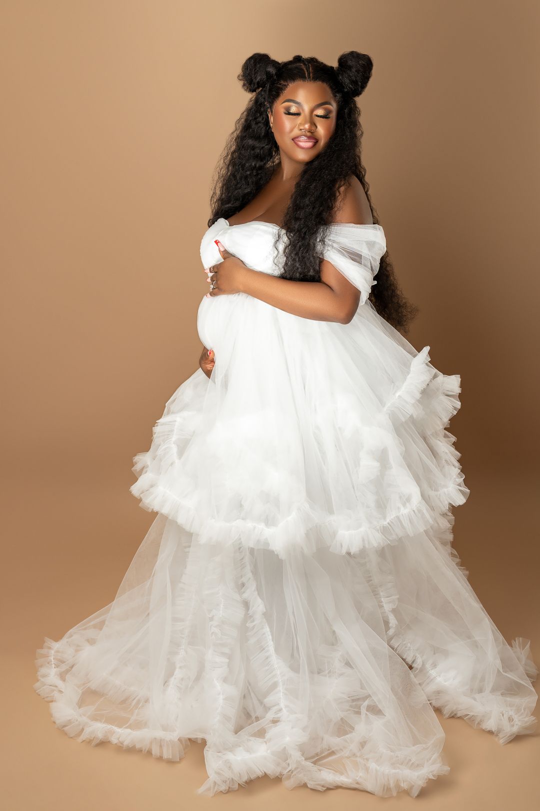 Graceful maternity portraits in white ruffled dress against brown background.