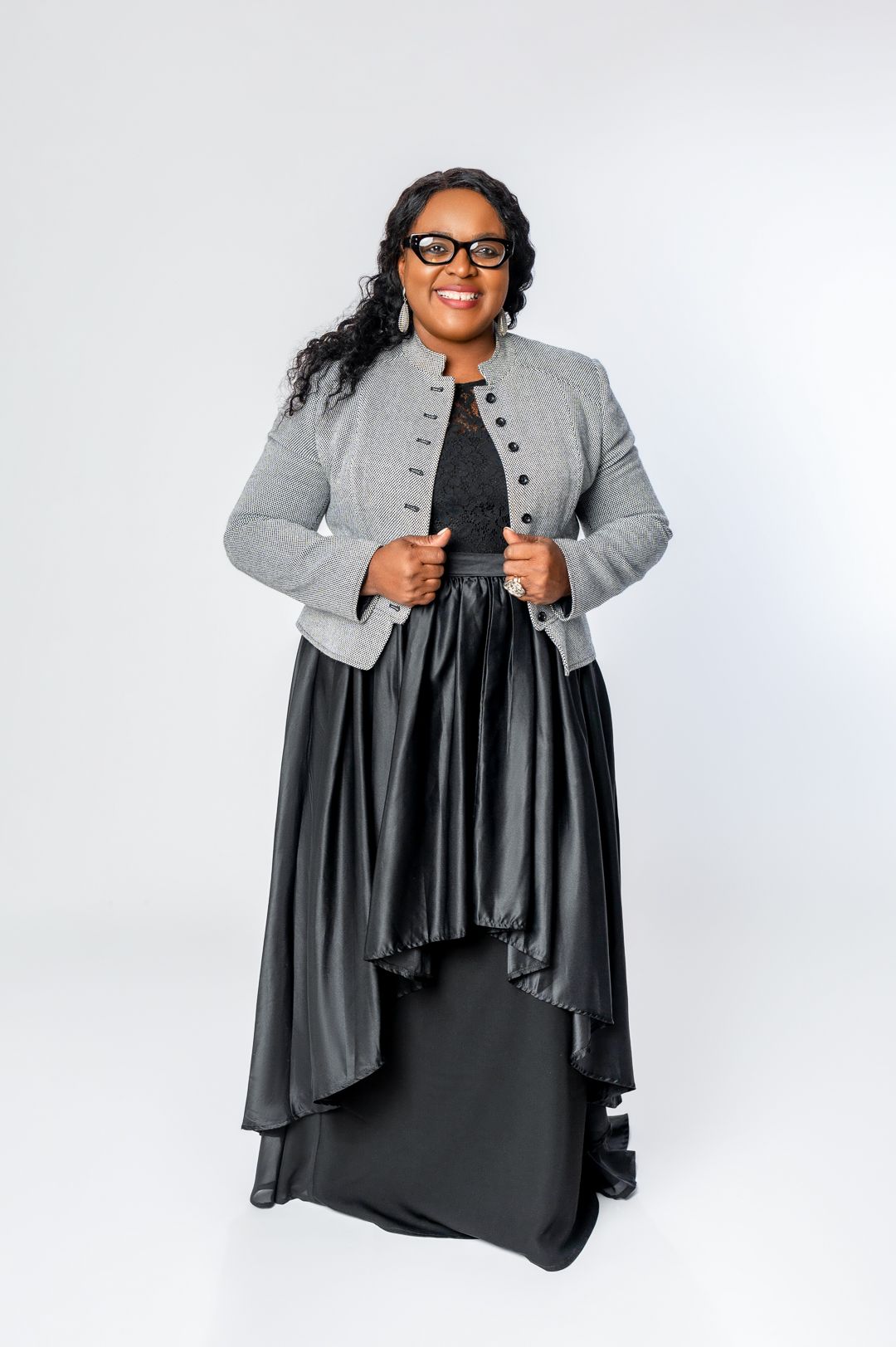 A portrait of someone wearing glasses in a gray military-style jacket and flowing black dress.