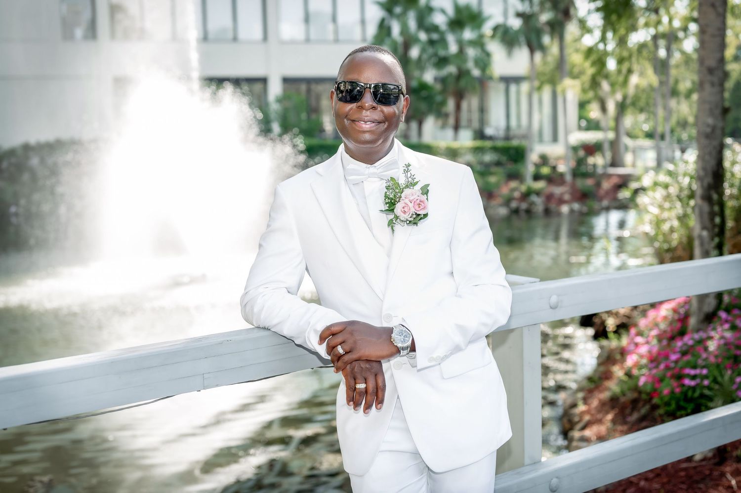 Groom in crisp white tuxedo poses confidently by fountain in garden setting.