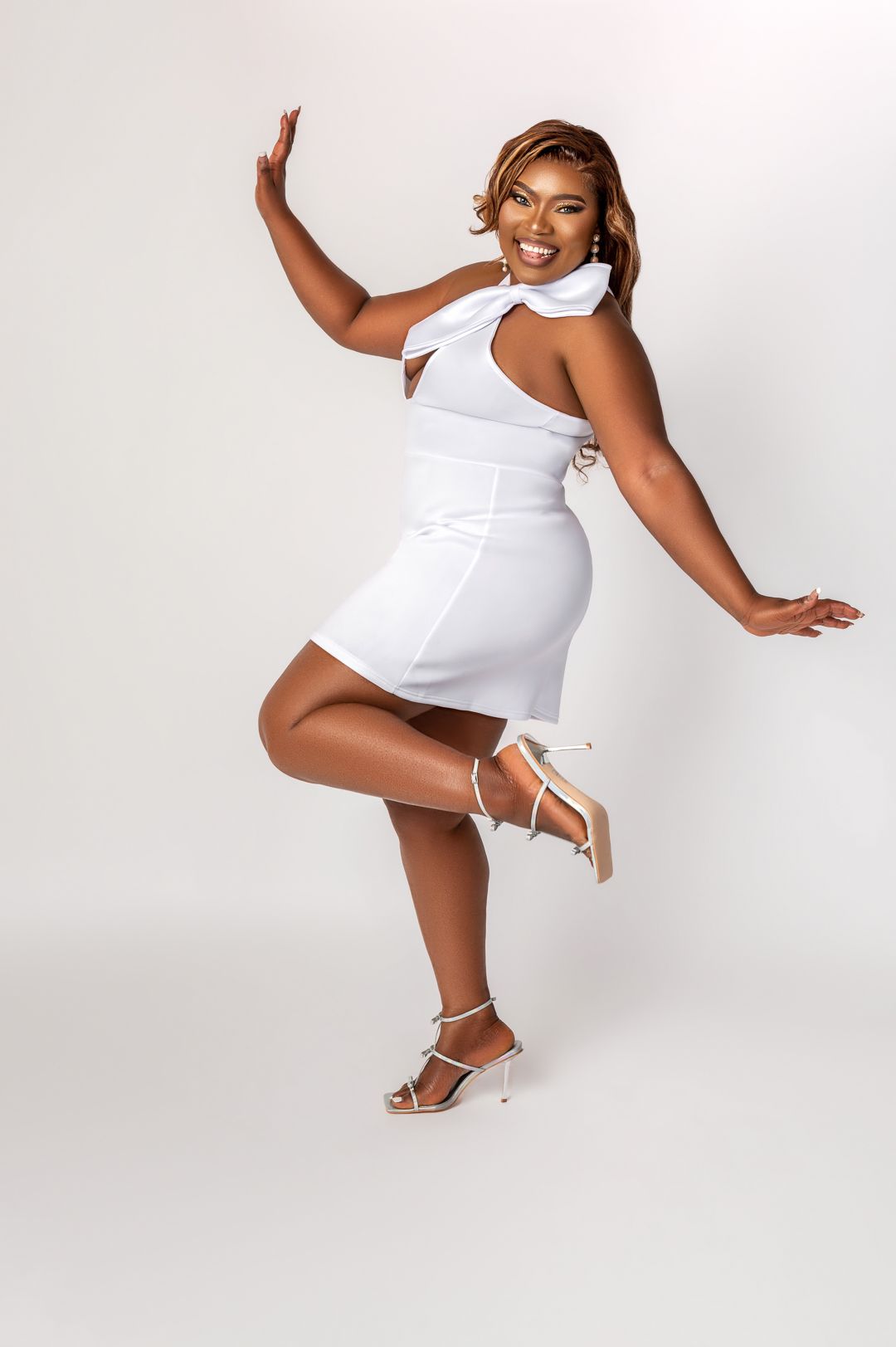 Joyful pose in white mini dress and strappy sandals against studio background.