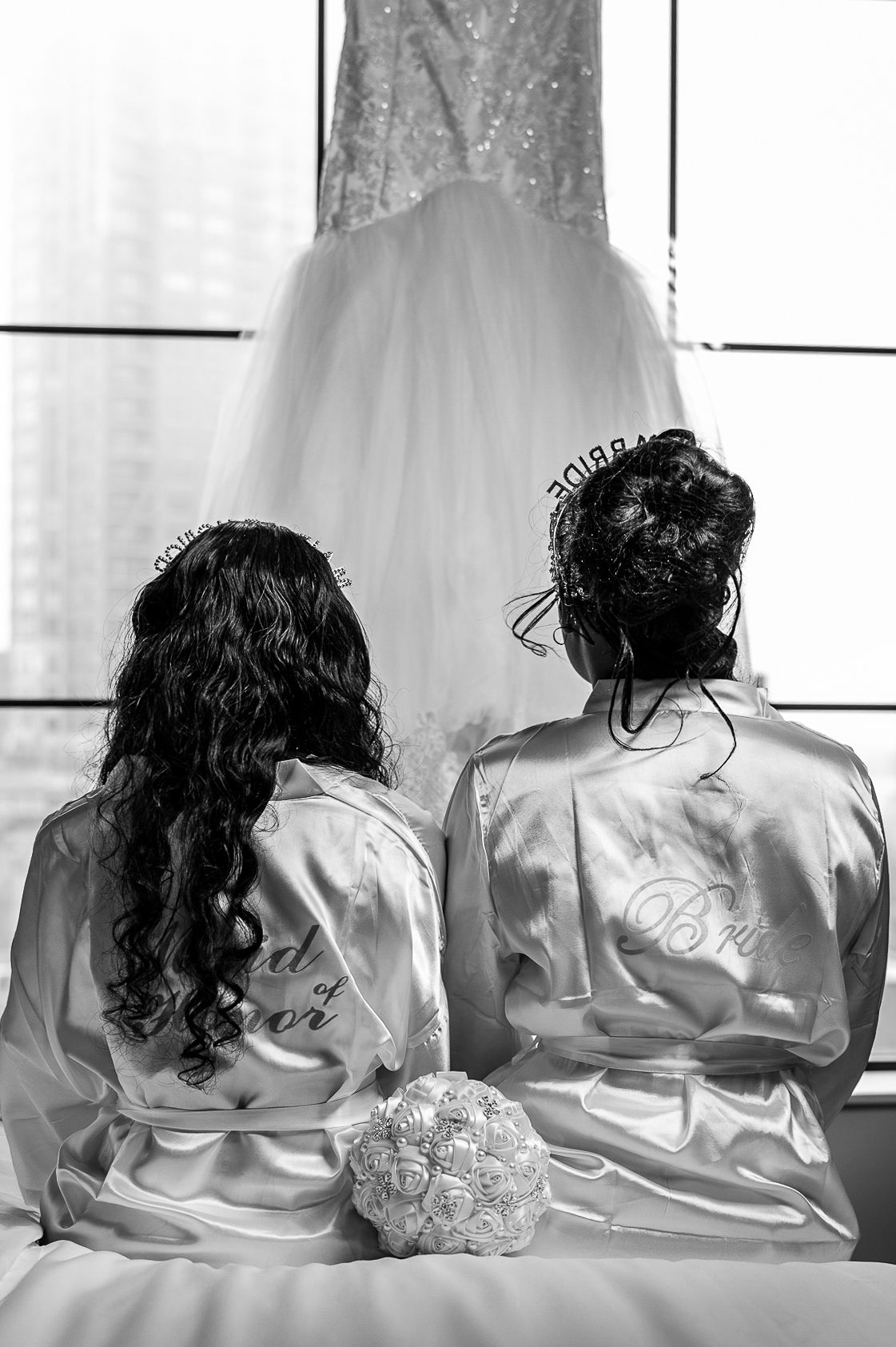 Back view of two bridesmaids in silk robes sitting beneath hanging wedding dress.