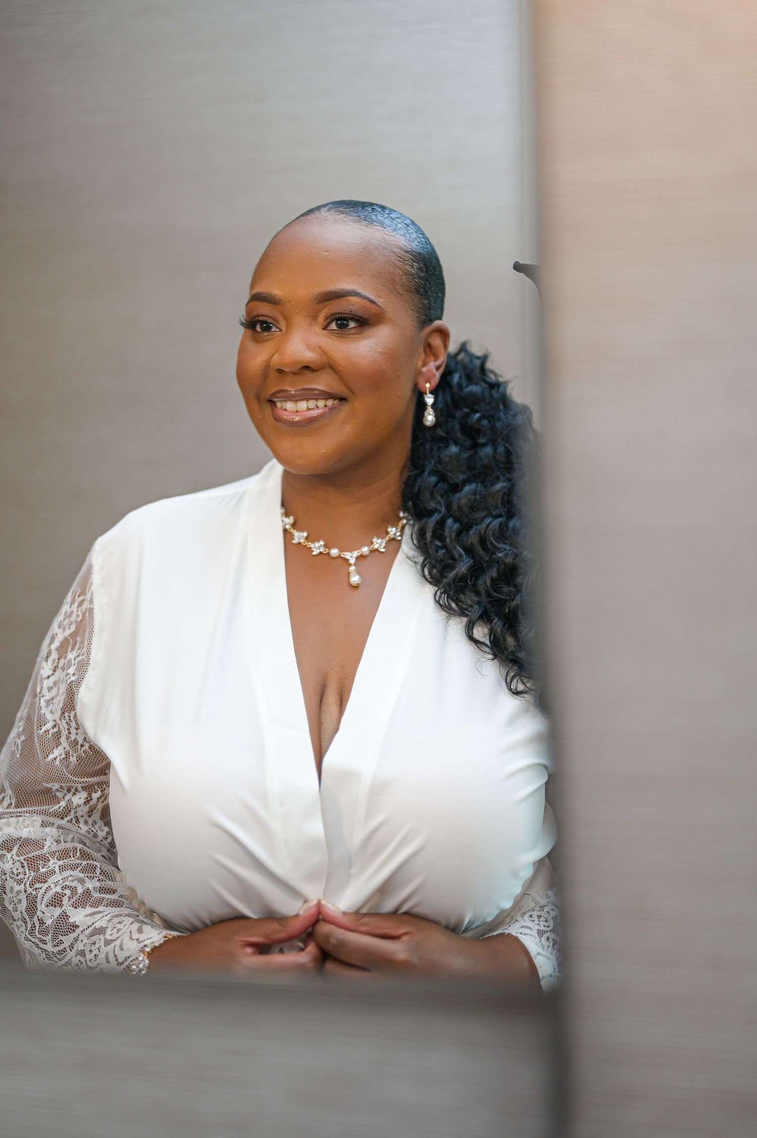 A woman in a white lace robe with elegant jewelry and styled hair poses during wedding preparations.