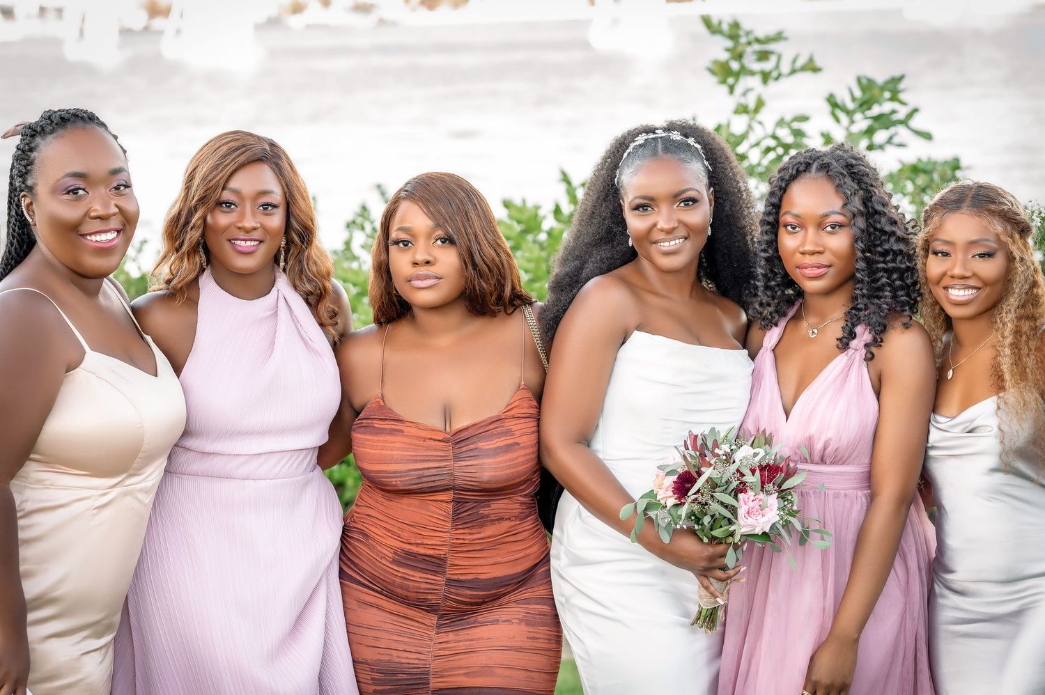 Bridesmaids in pink and neutral dresses pose with flowers at outdoor wedding.