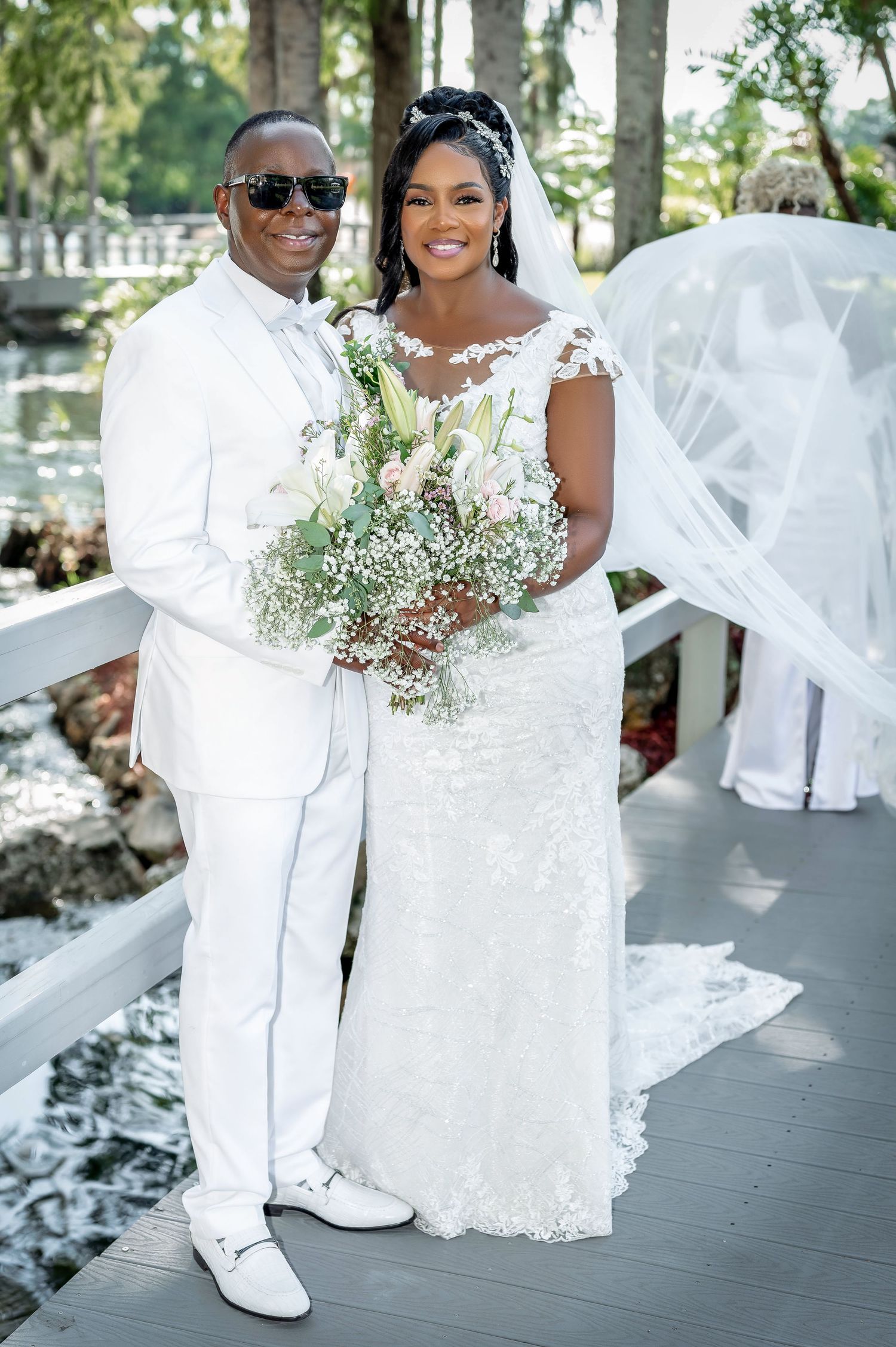 Couple in elegant white attire poses on deck with floral bouquet during outdoor wedding.