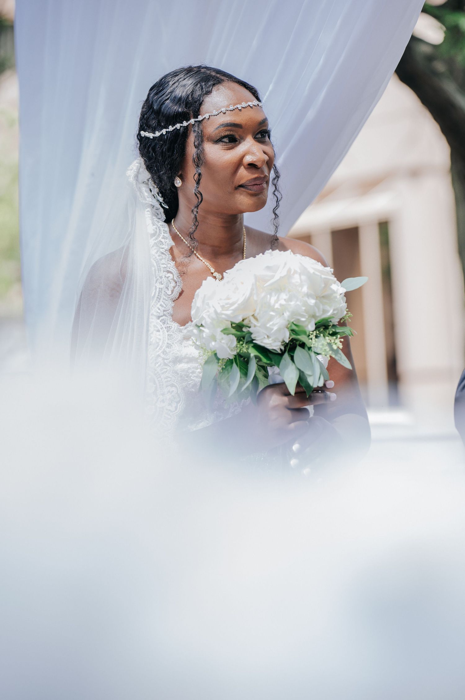 Close-up portrait through white veil of bride holding white bouquet with delicate hair accessories.