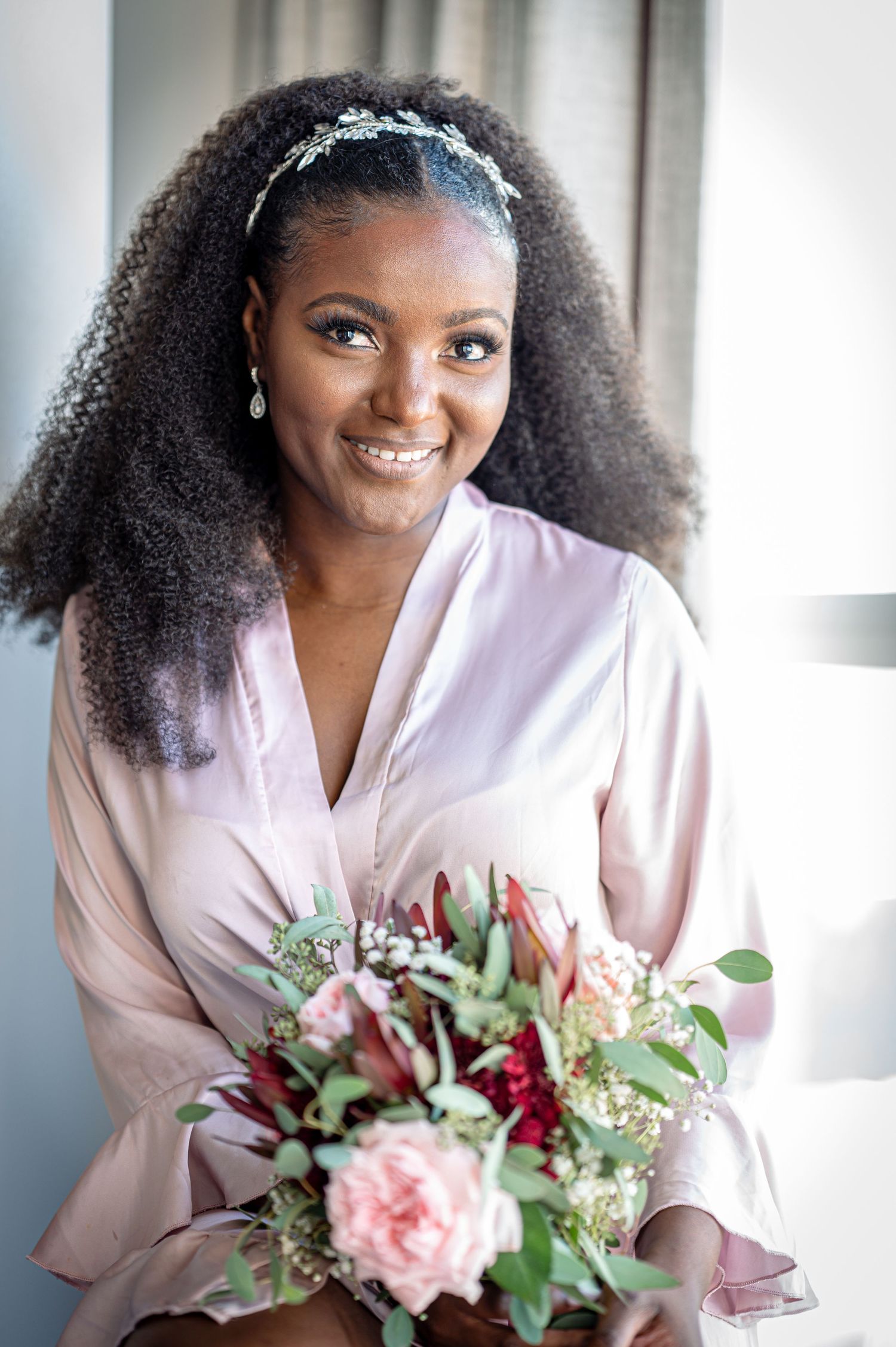 A beaming bride in a pink silk robe holds a blush and burgundy floral bouquet during wedding preparations.