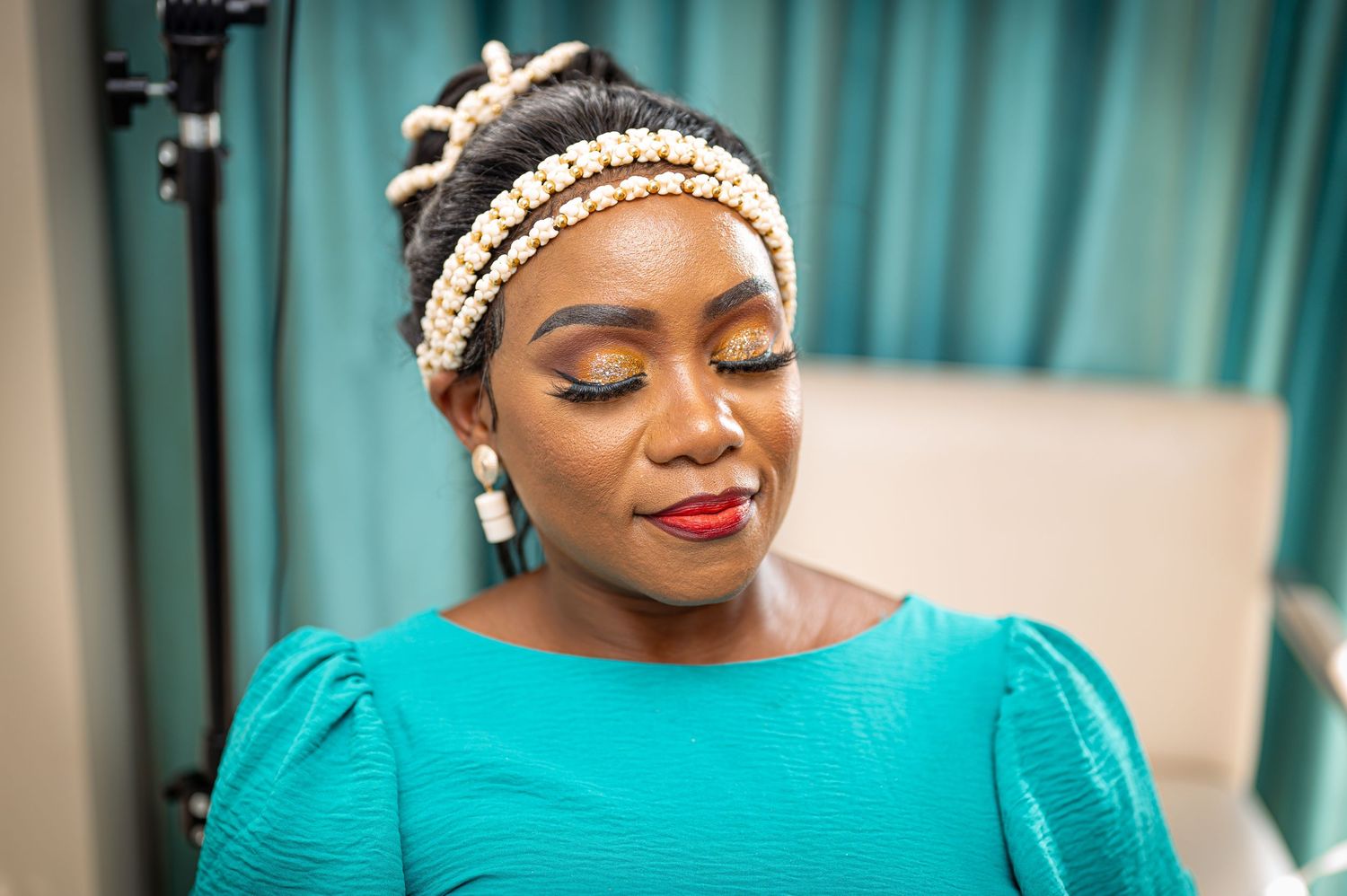 A person wearing a teal dress and pearl headpiece with elegant makeup against a blue backdrop.