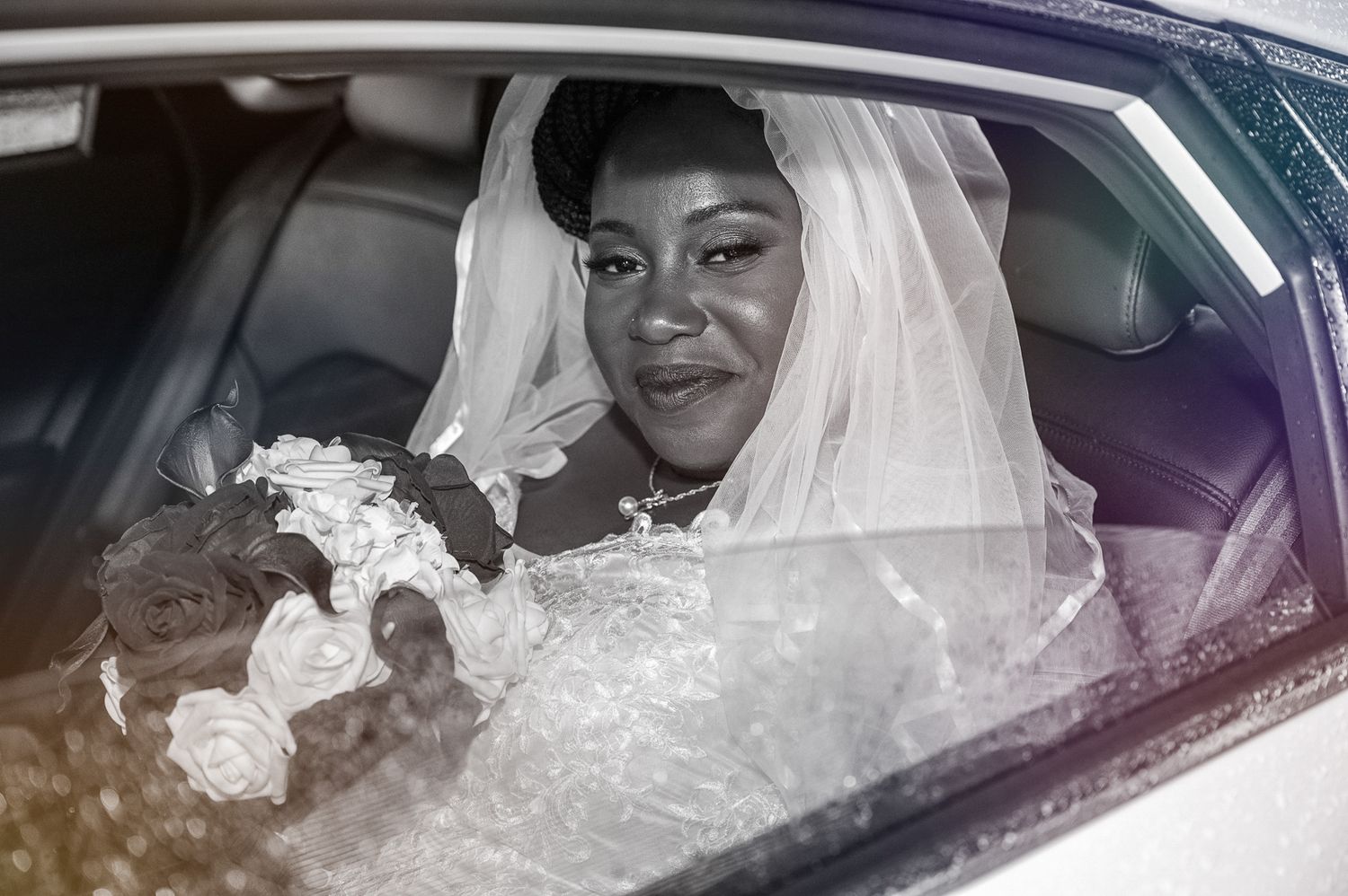 A bride smiles radiantly through a car window, wearing a veil and white dress.