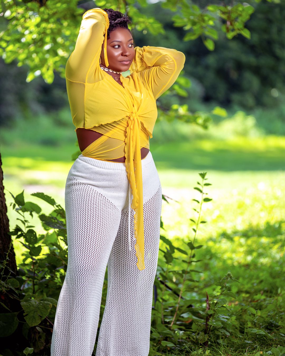 Fashion photoshoot in a sunny garden setting featuring white pants and yellow top against lush greenery.