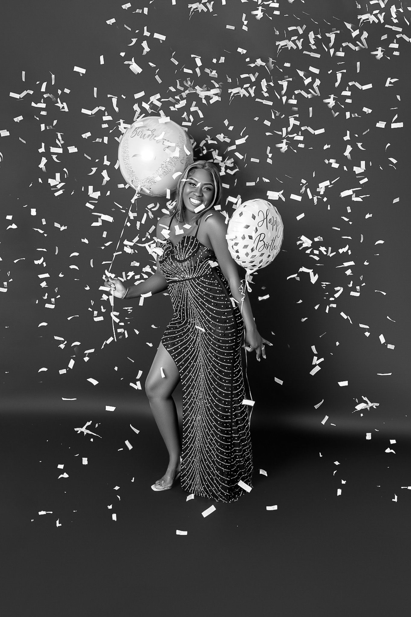 Figure in sparkly dress celebrates with balloons and confetti in black and white photo.