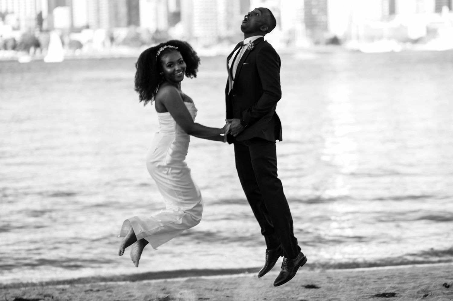 Black and white photo of a couple jumping together on a beach during a wedding photoshoot.