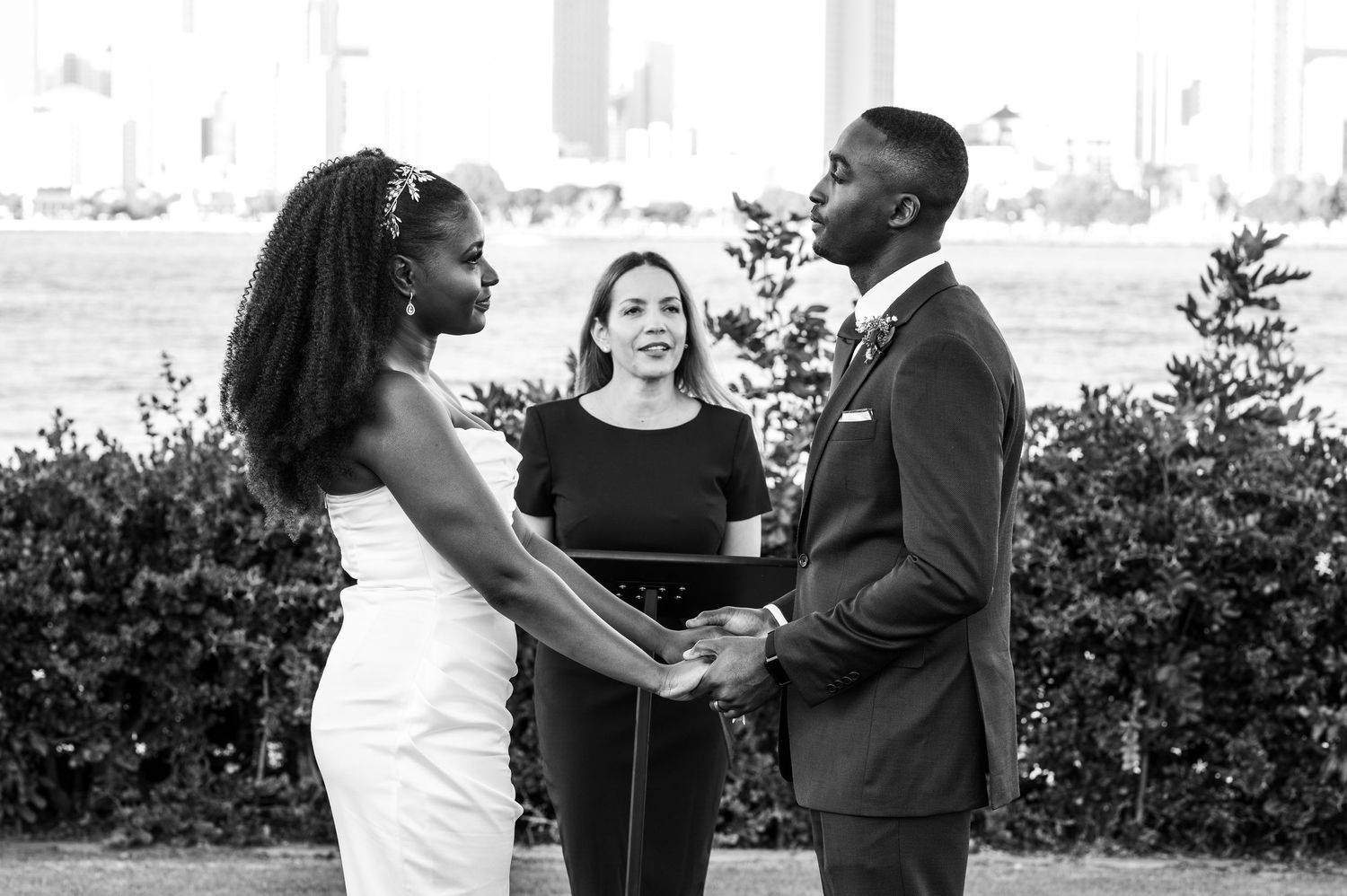 Black and white photo of outdoor wedding ceremony with couple holding hands and officiant.