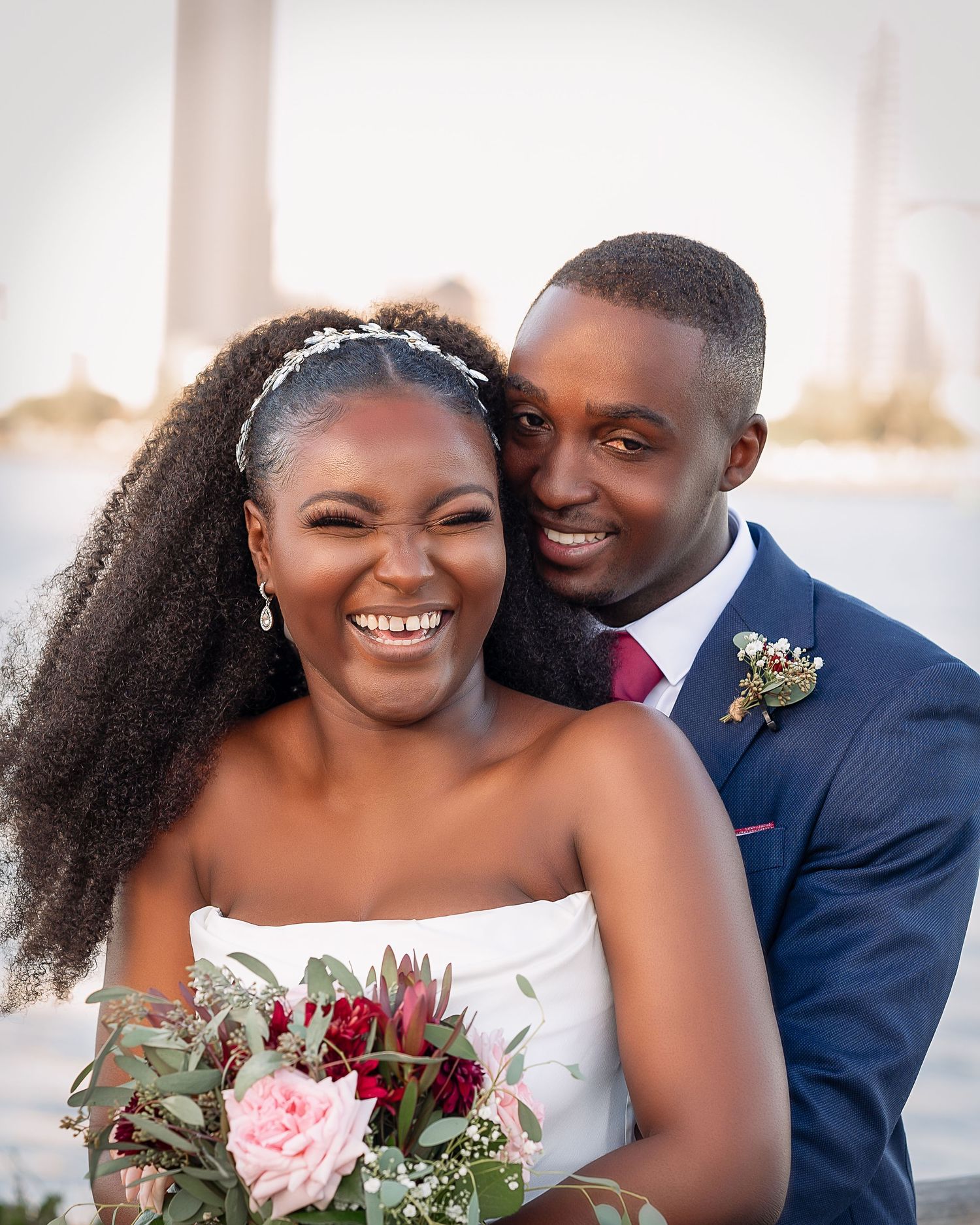 Romantic outdoor wedding portrait of newlyweds smiling with colorful bouquet by waterfront.