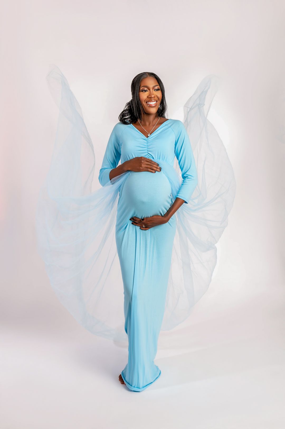 Expectant mother in light blue dress poses with ethereal white fabric wings.