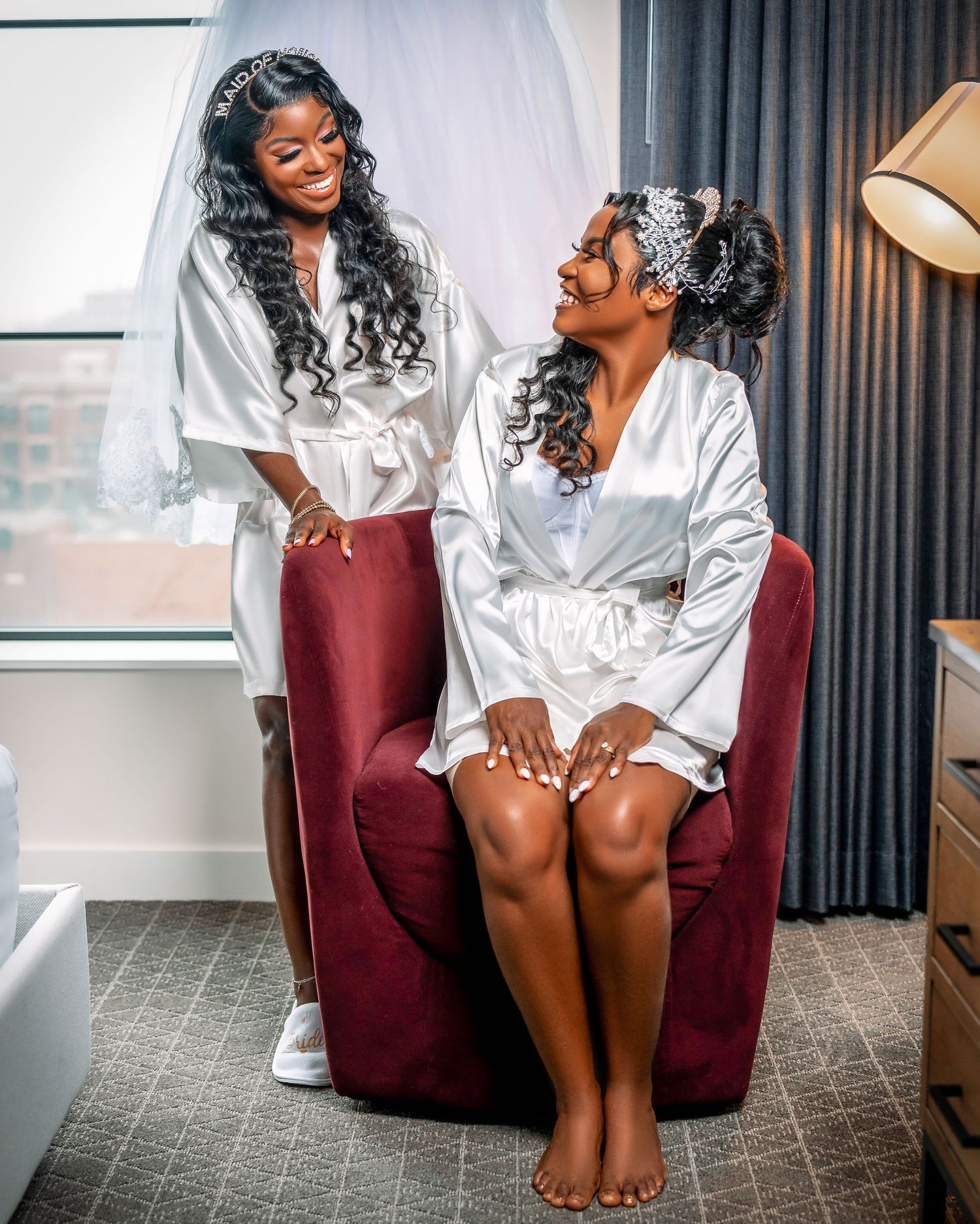 Two women in white silk robes sharing a joyful moment on a burgundy chair during wedding preparations.