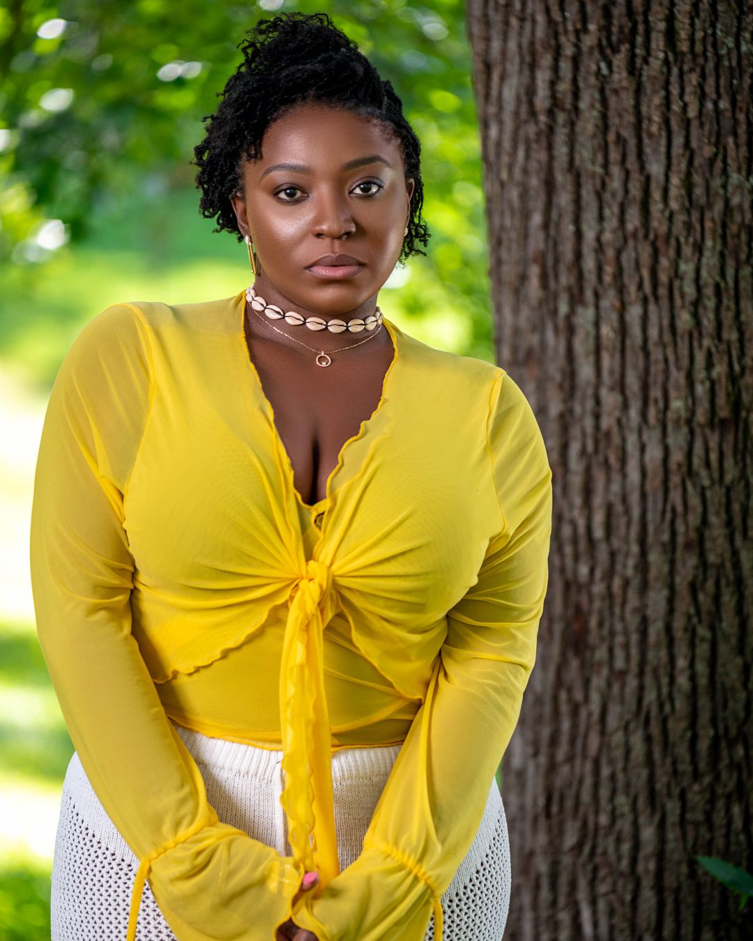 Outdoor portrait in yellow knotted top and white skirt against green foliage background.