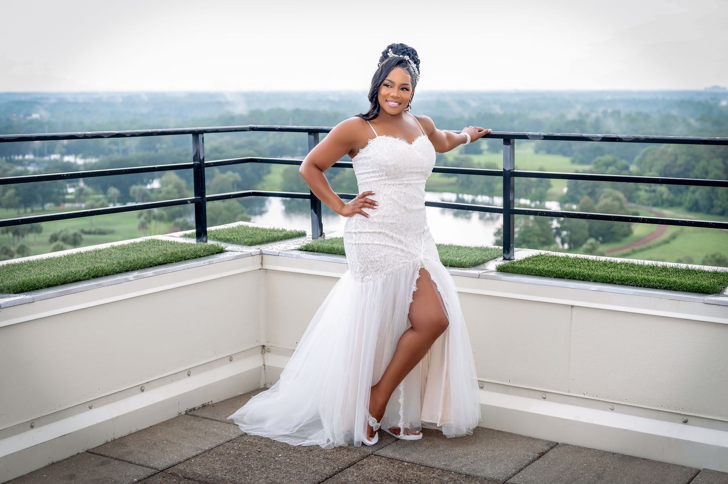A person in a white wedding gown with high slit poses on an outdoor balcony with scenic view.