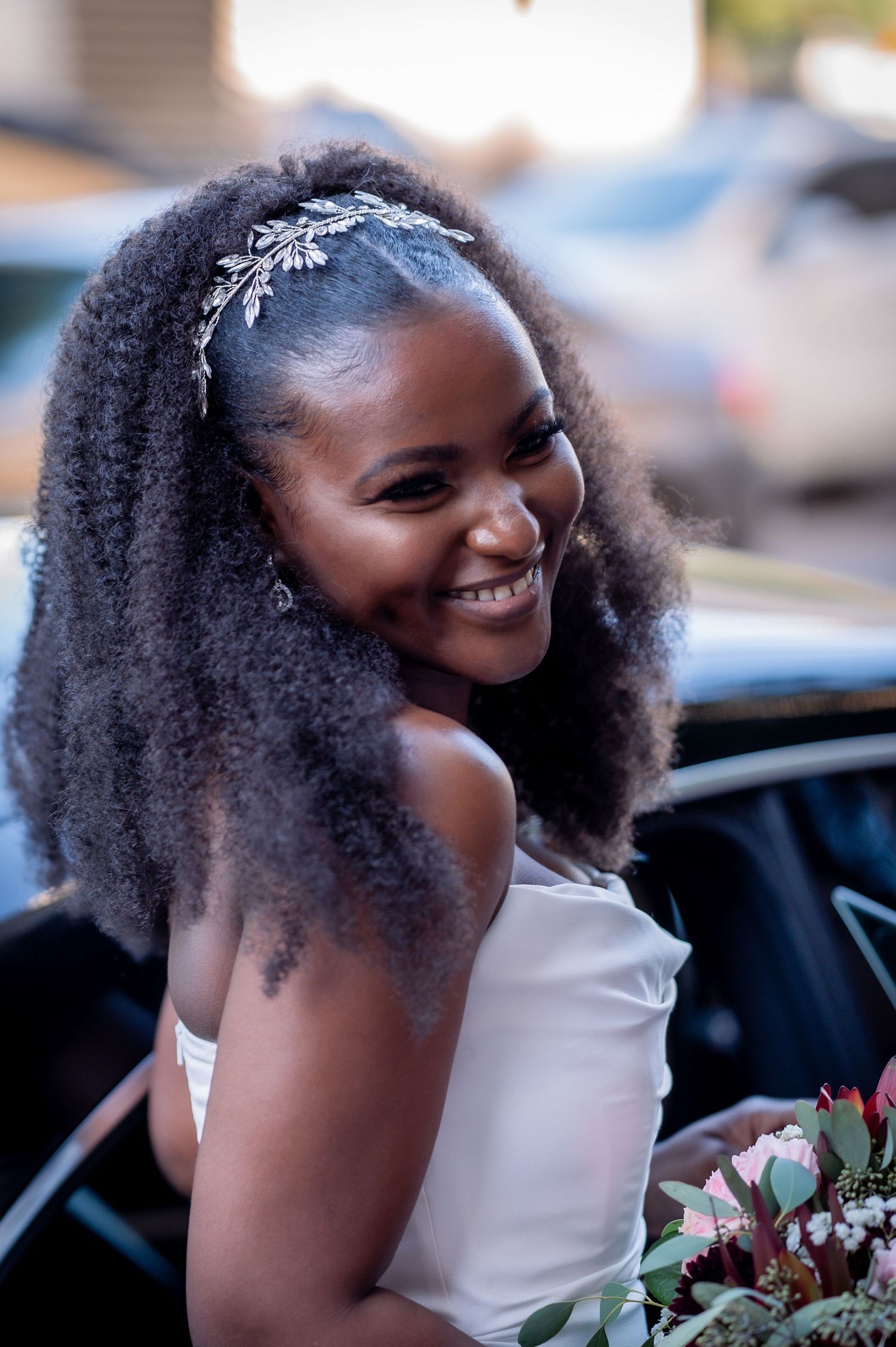 Smiling bride with natural curls wearing crystal headpiece holds bouquet.