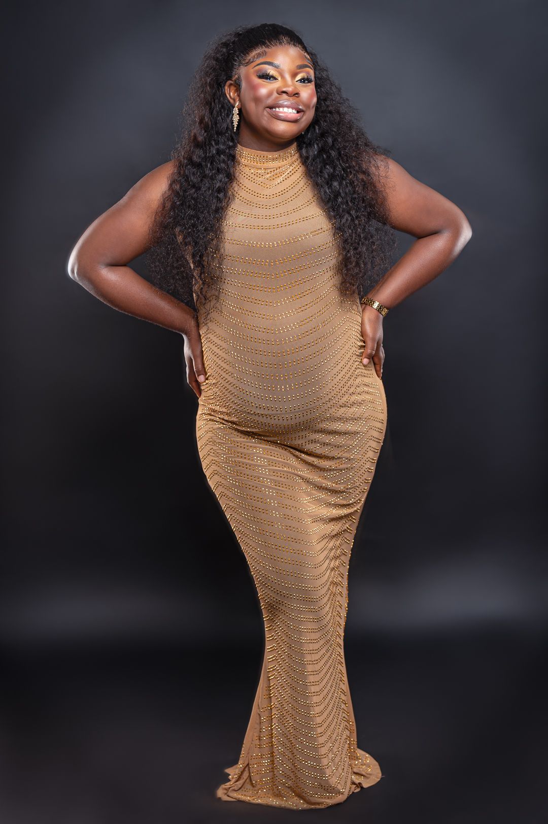 A person in a fitted tan gown poses confidently against dark background.