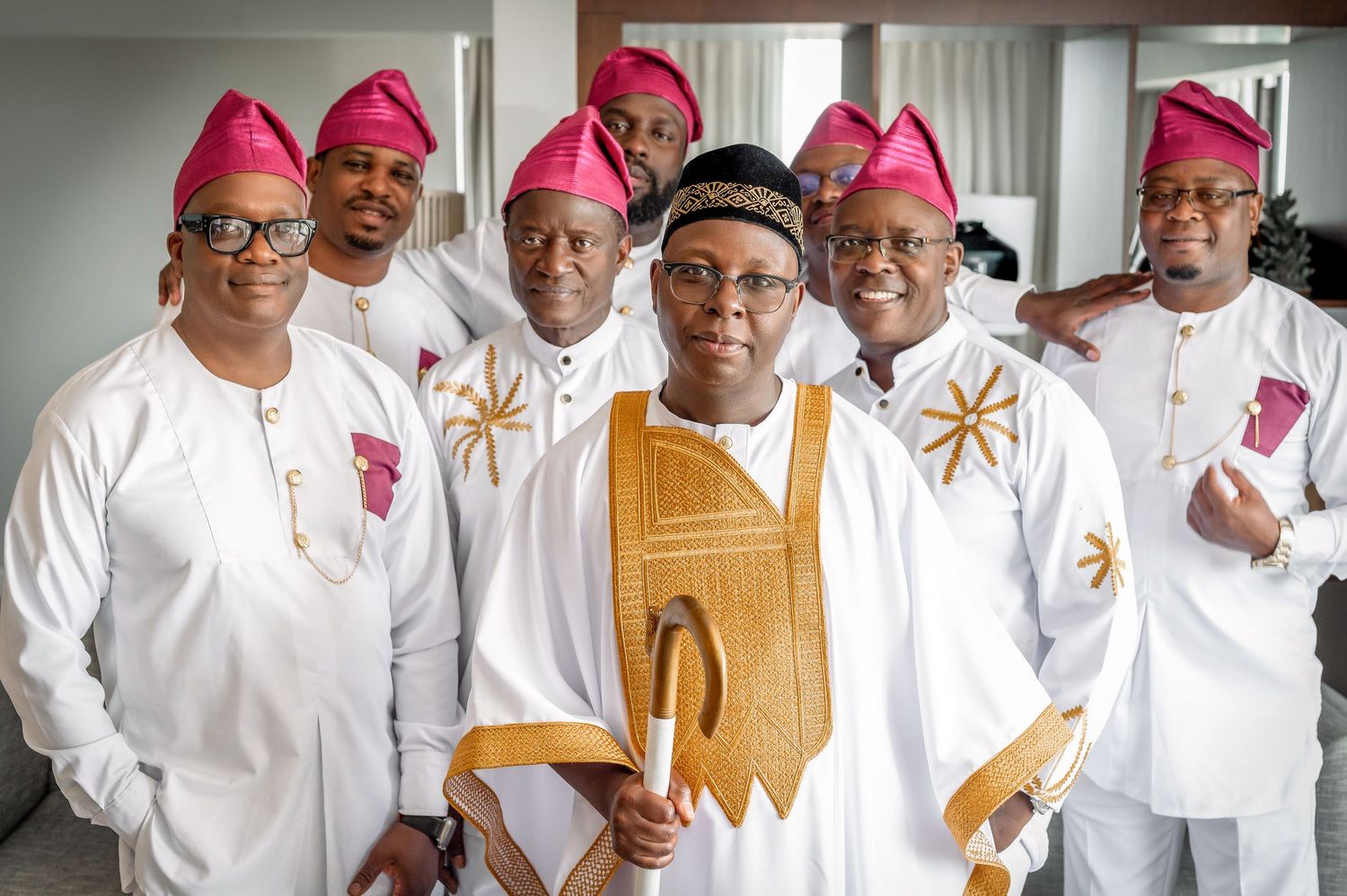 Traditional wedding party in white and gold attire with pink ceremonial caps.