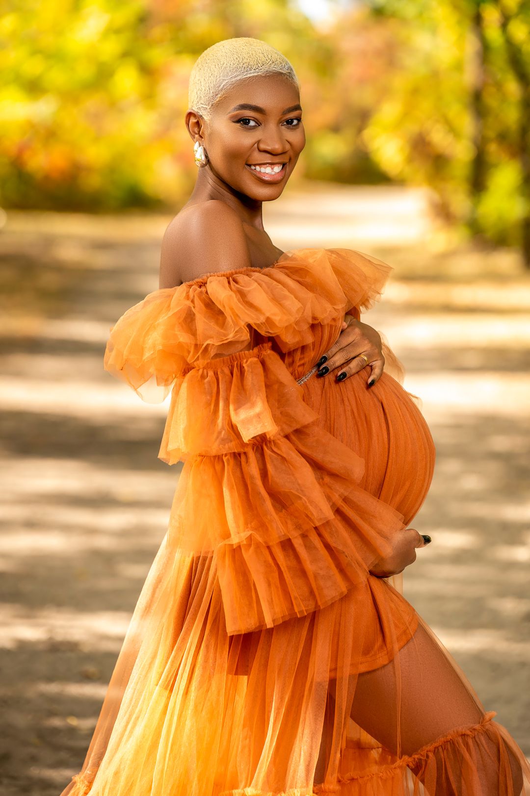 Expectant mother in flowing orange tulle gown poses outdoors during autumn maternity photoshoot.