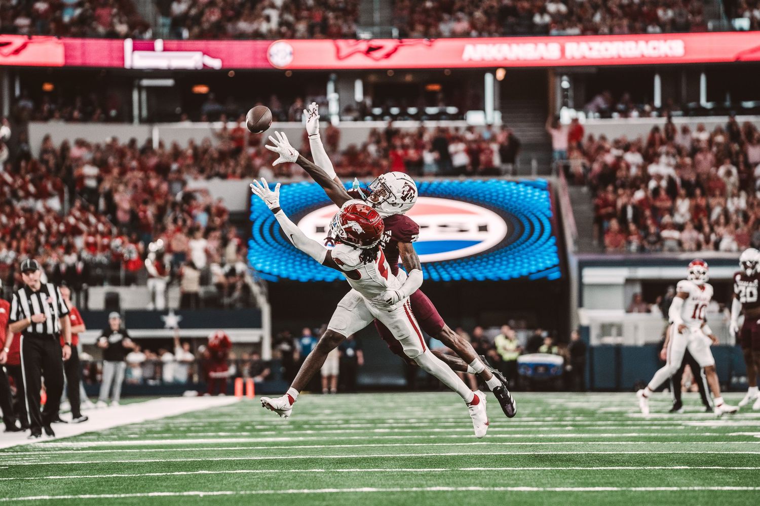 Two football players leap high to compete for a catch during a game in a packed stadium.