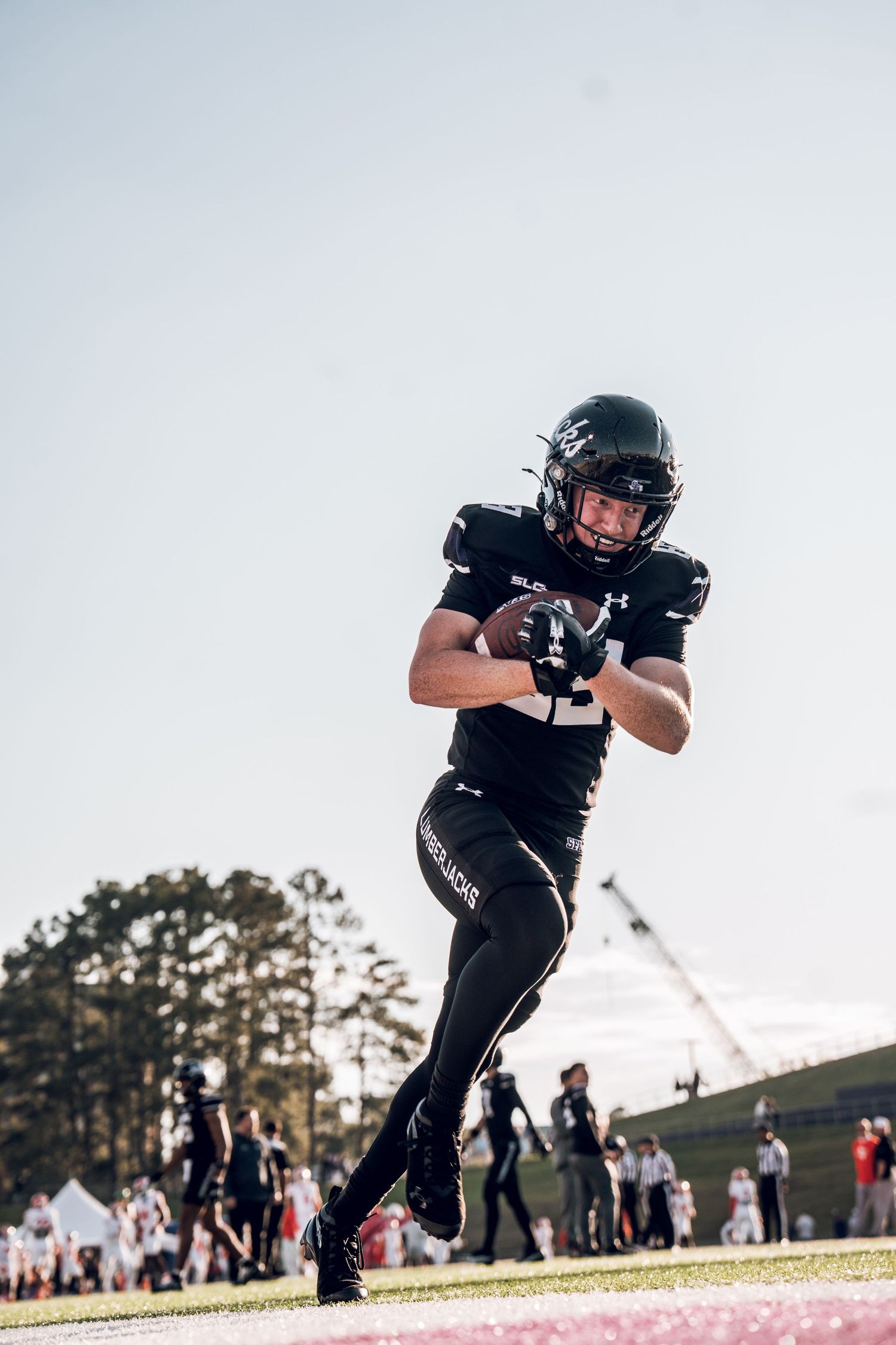 Football player in black uniform runs across field during game with spectators and trees in background.