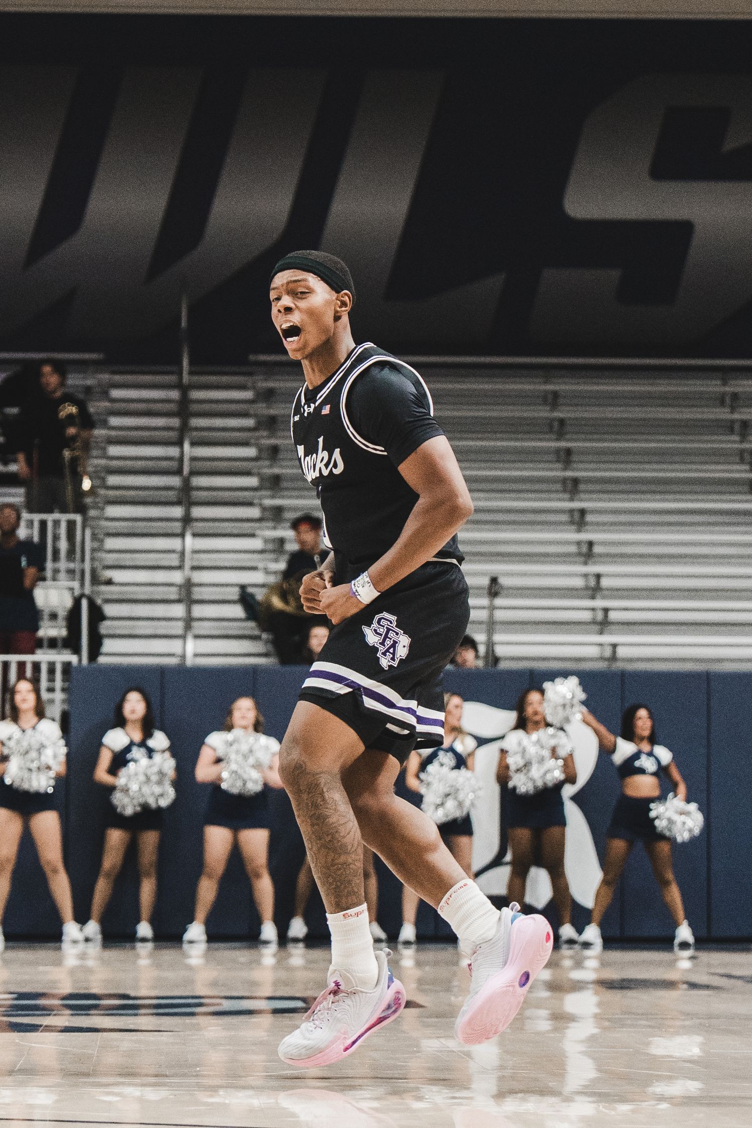 Basketball player wearing black jersey performs dynamic move on court with cheerleaders in background.