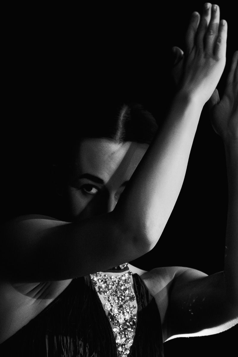 Black and white portrait of a dancer in sequined dress with arms raised gracefully against dark background.
