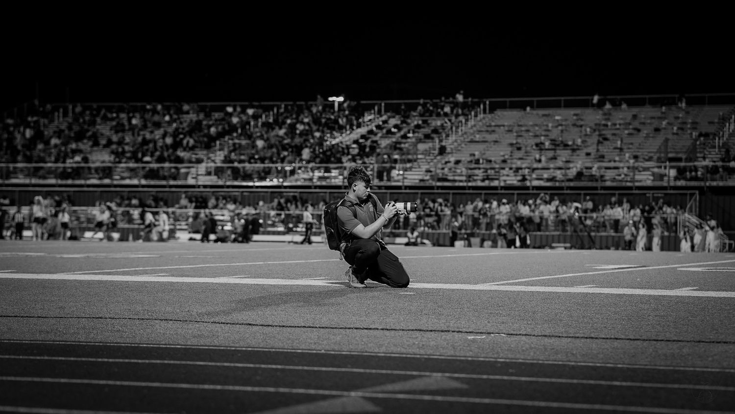 Black and white image of motorcycle racing on a track with stadium crowd in background.