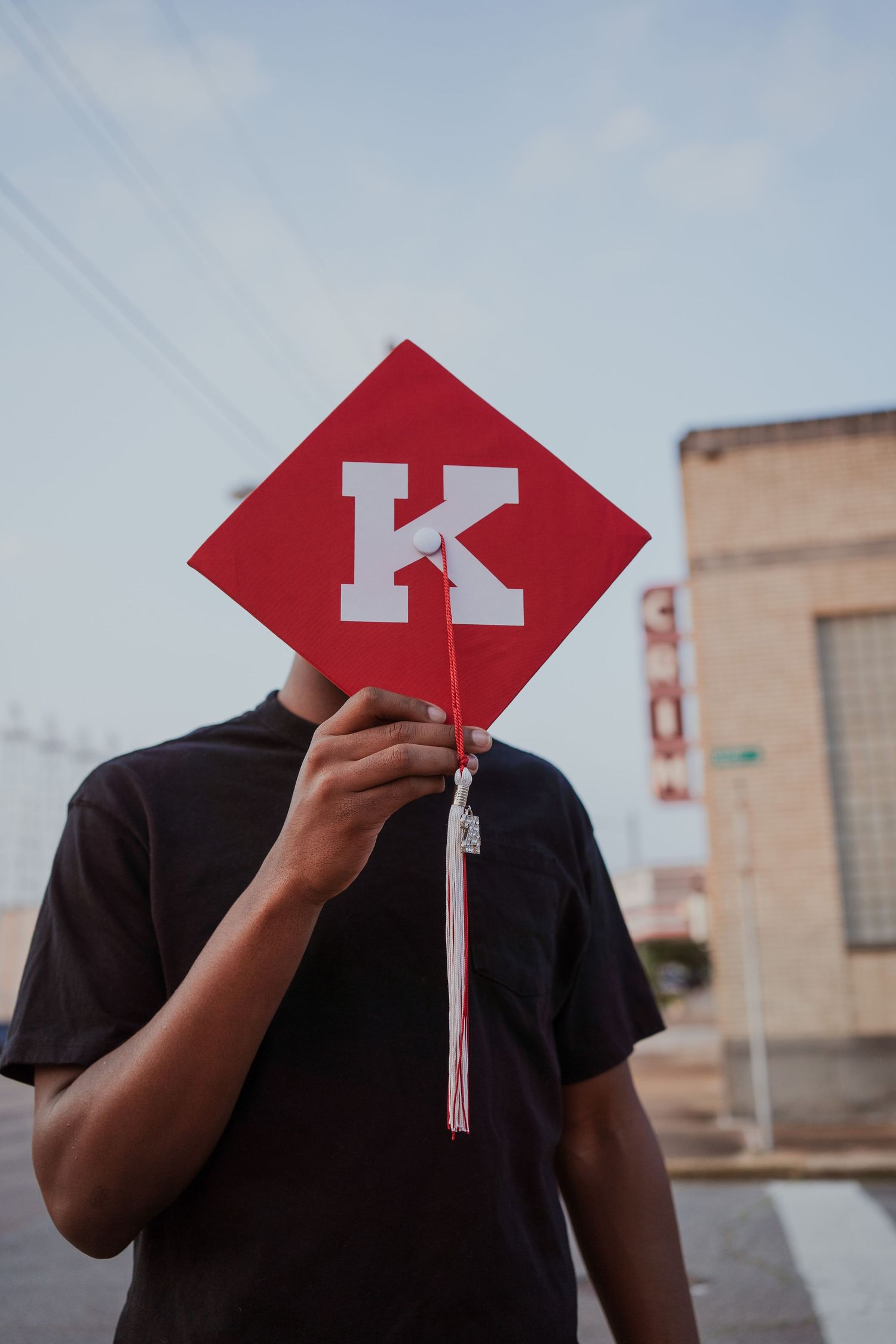 Graduate holds up red graduation cap with letter K against urban backdrop.