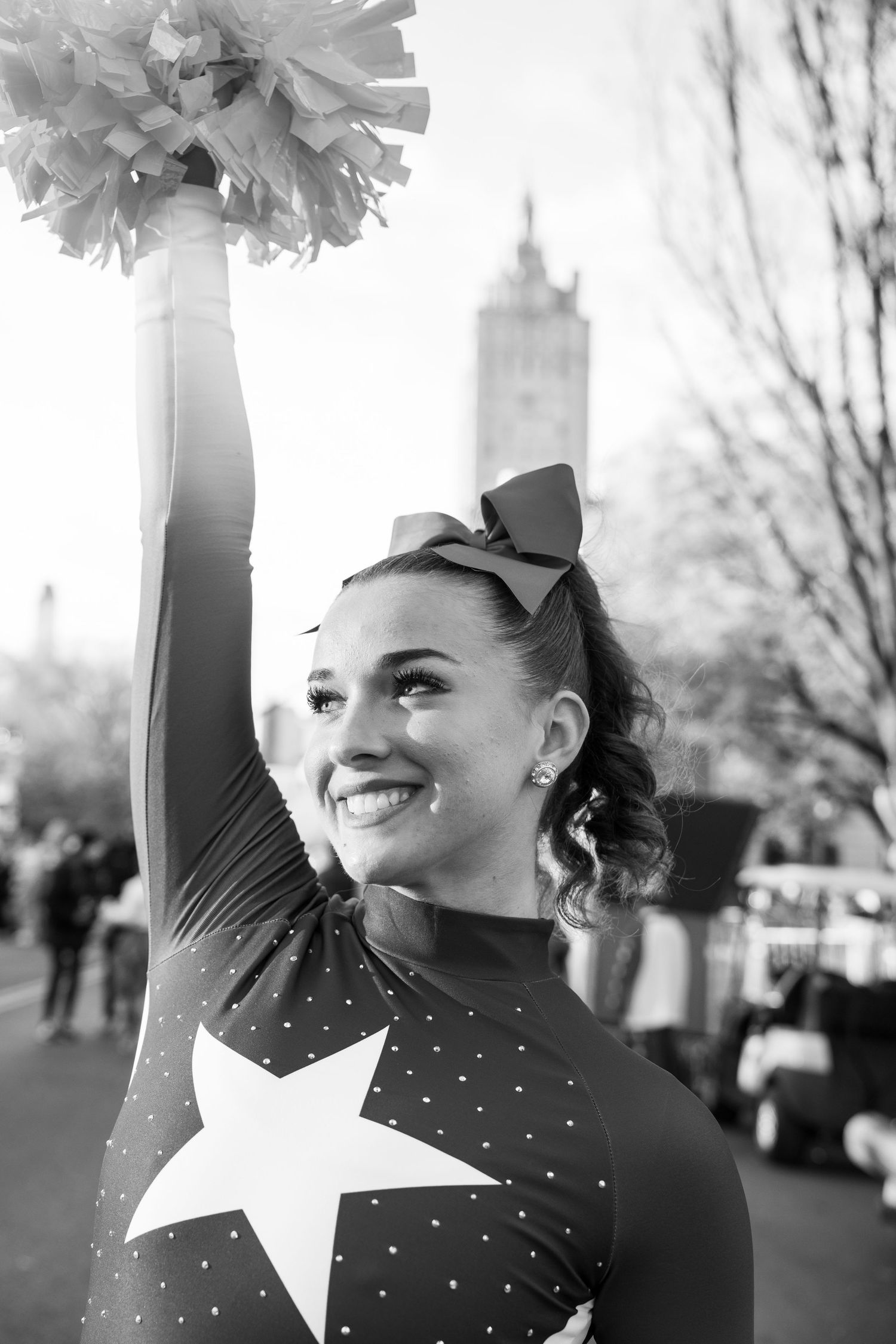 Cheerleader in black and white sparkly uniform with white bow performs energetic routine outdoors.