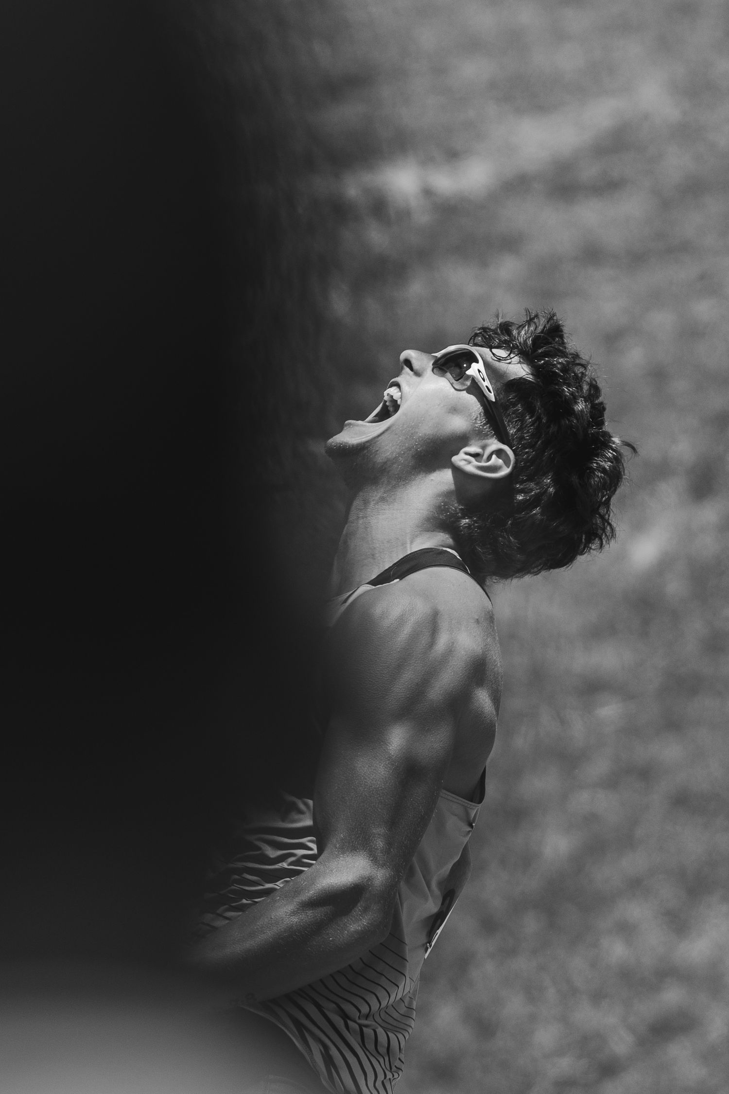 Black and white photo of a muscular man with curly hair, head tilted back, mouth open, wearing sunglasses outdoors.