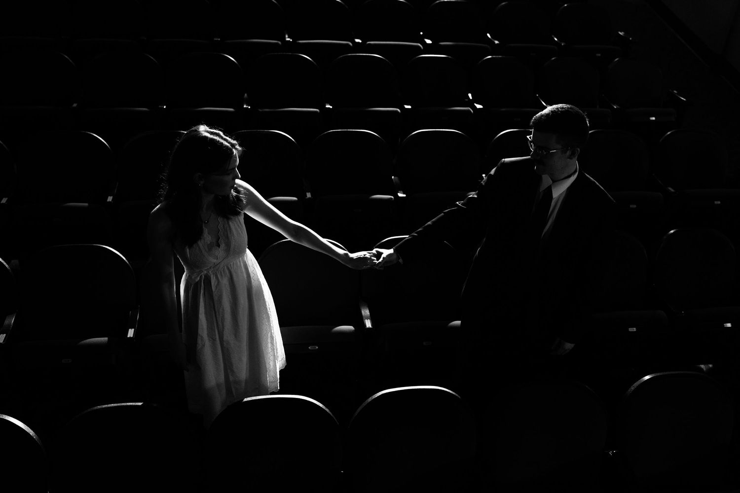 Black and white photo of a couple holding hands in a dark theater aisle, dressed formally.
