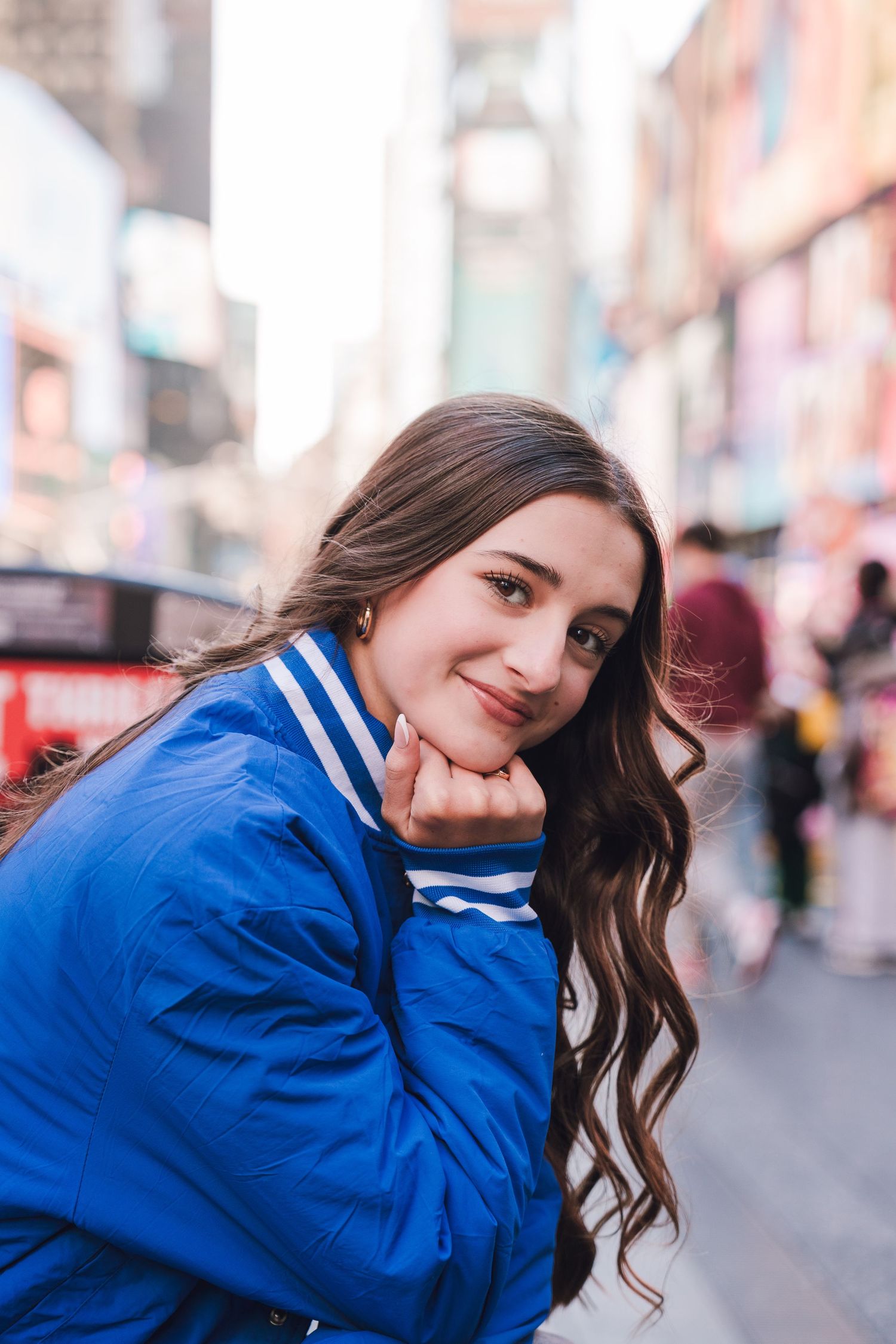 Young woman in blue varsity jacket smiling in Times Square NYC, with blurred city lights and buildings in background.