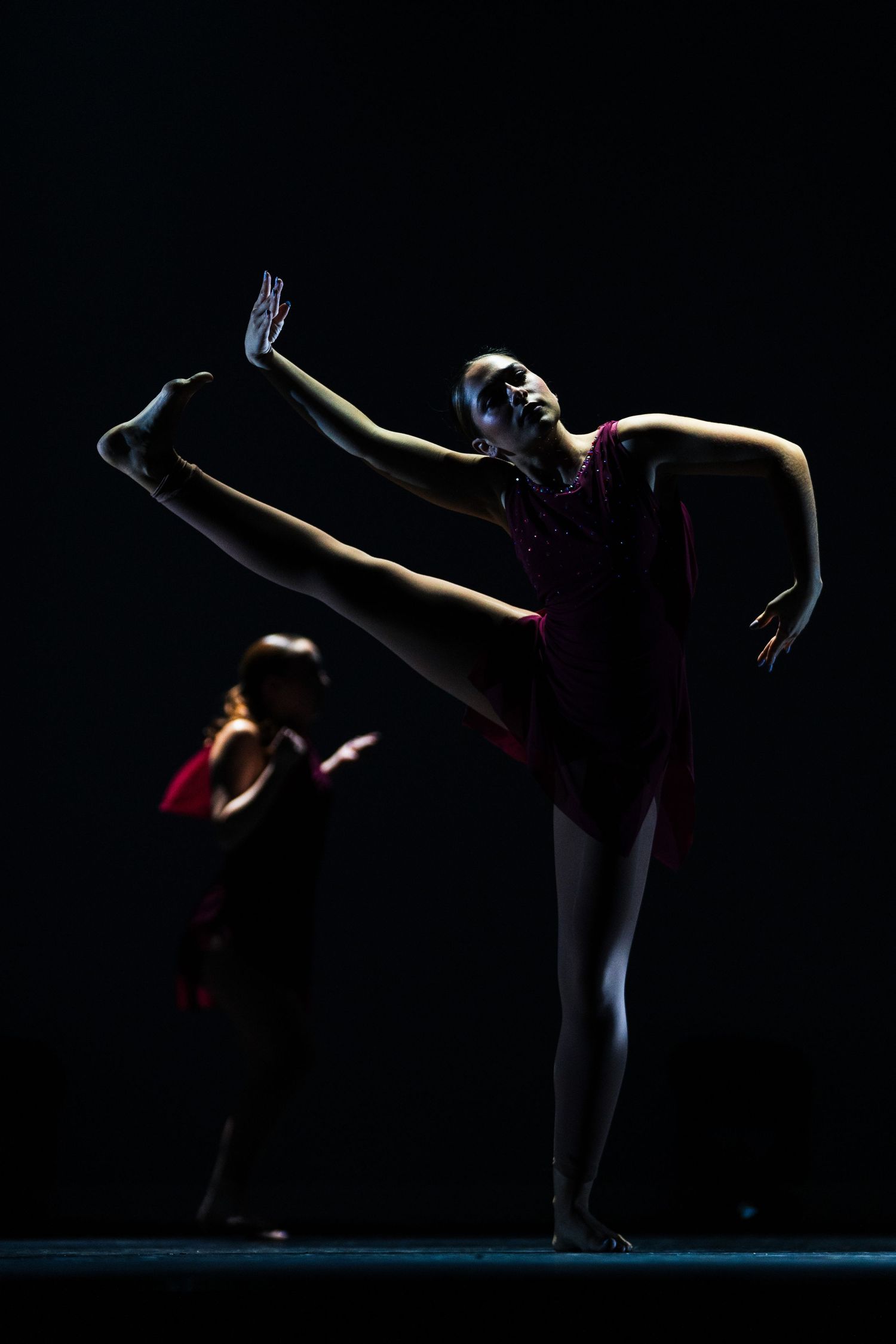 Two ballet dancers in red costumes perform gracefully on a dark stage, one executing a high leg extension.