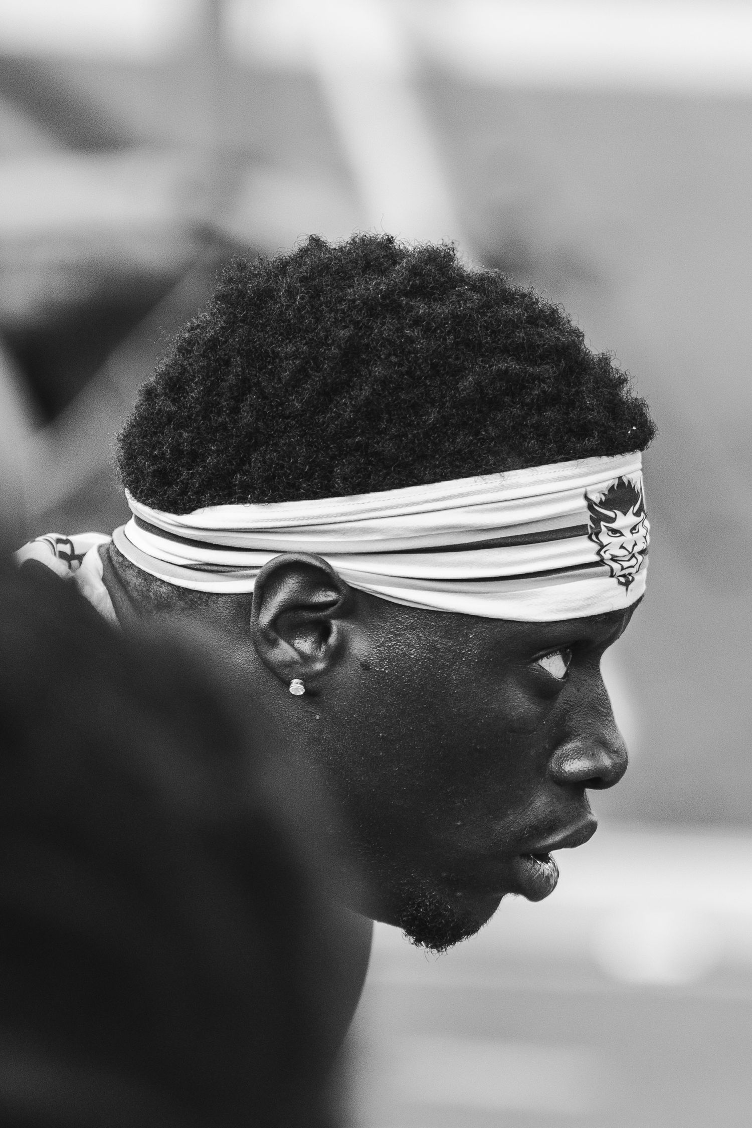 Black and white profile portrait of a young Black man wearing a white headband with a logo, featuring natural hair and a stud earring.