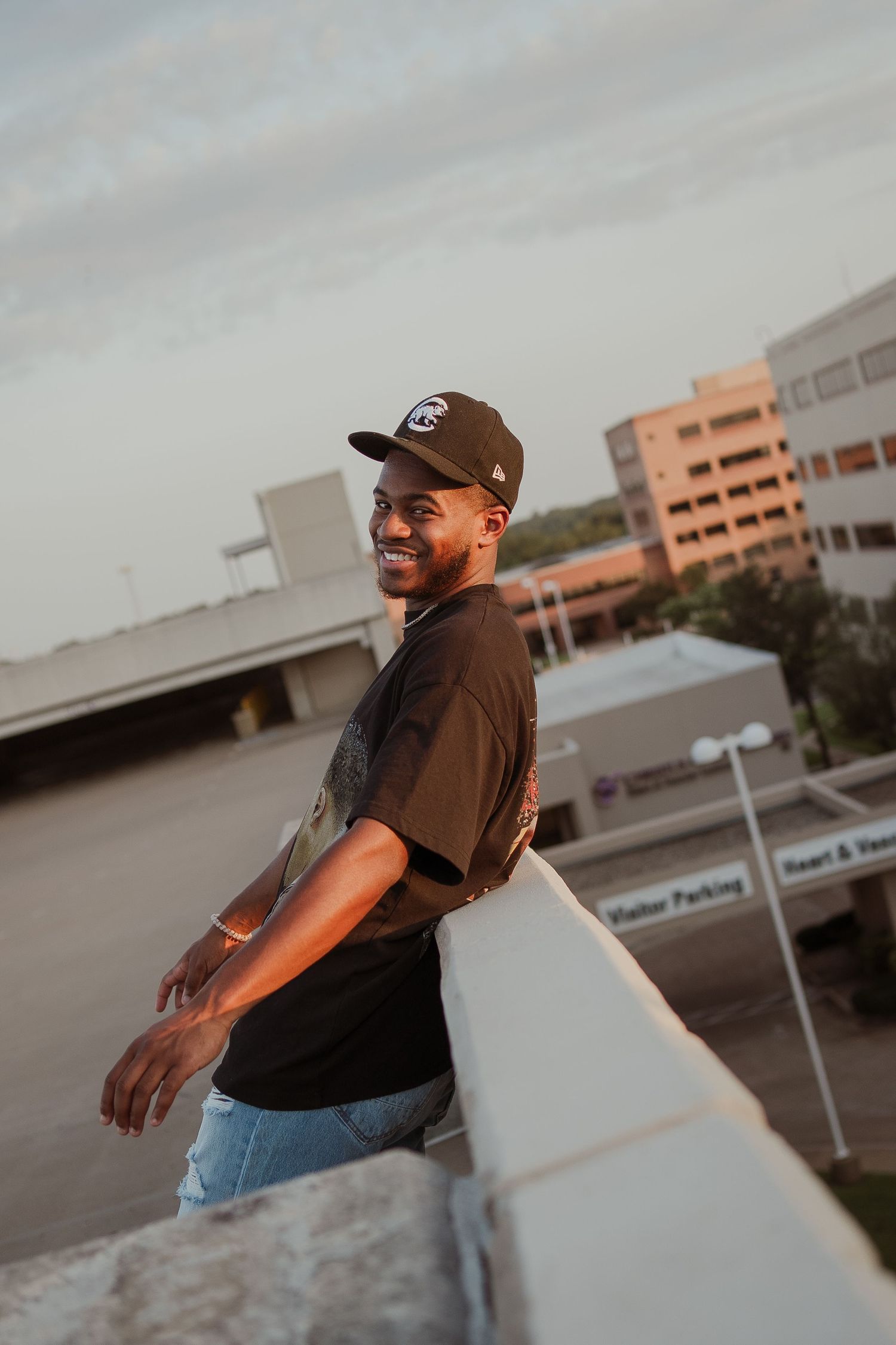 Person in baseball cap and casual wear poses on rooftop during golden hour.