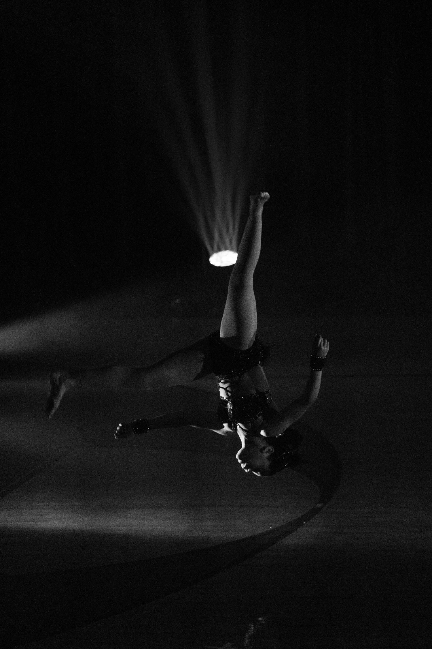 Dramatic black and white photograph of a dancer performing an upside-down aerial move with a spotlight in darkness.