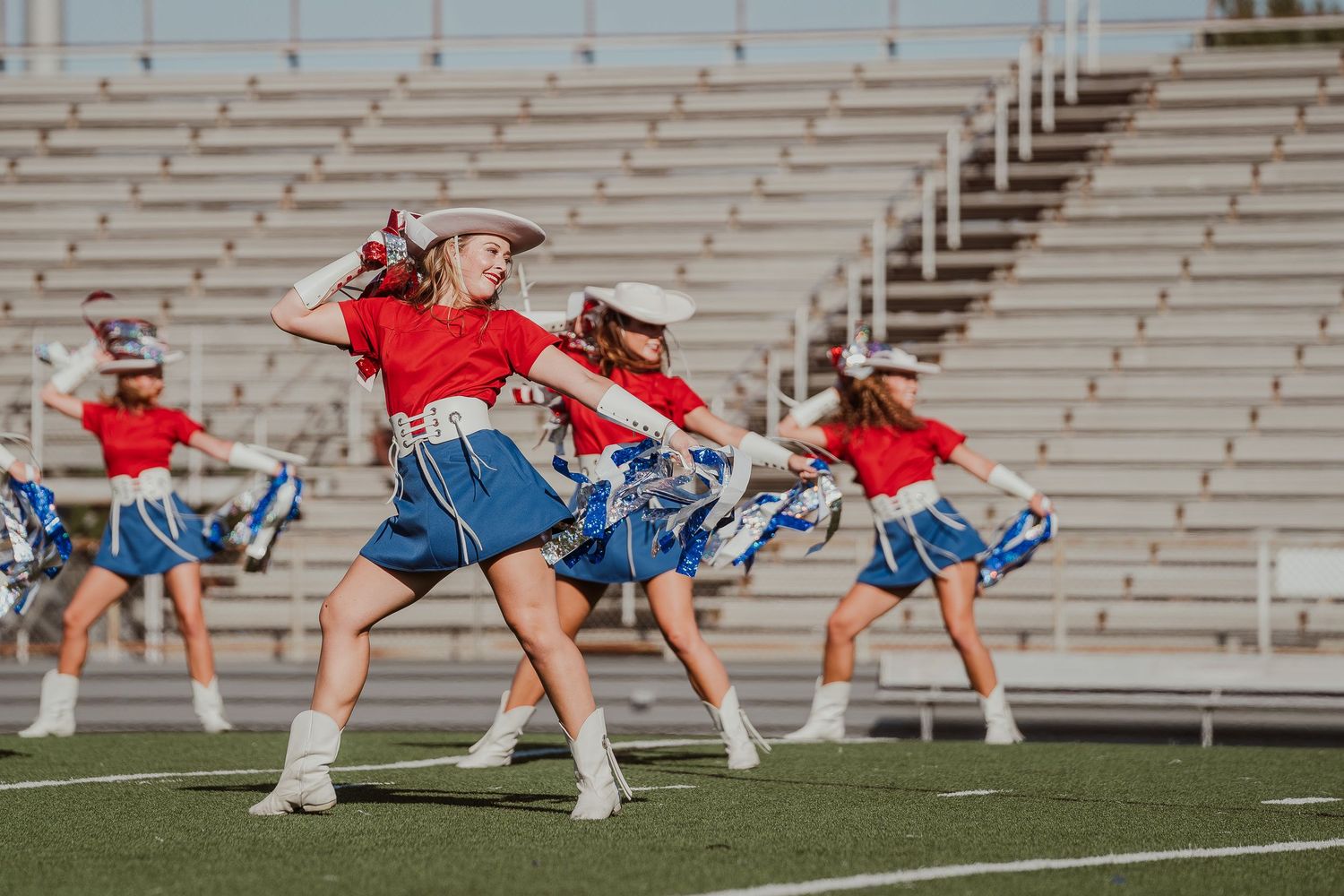 Dance team performers in red tops and blue skirts practice a synchronized routine on a football field with stadium bleachers.